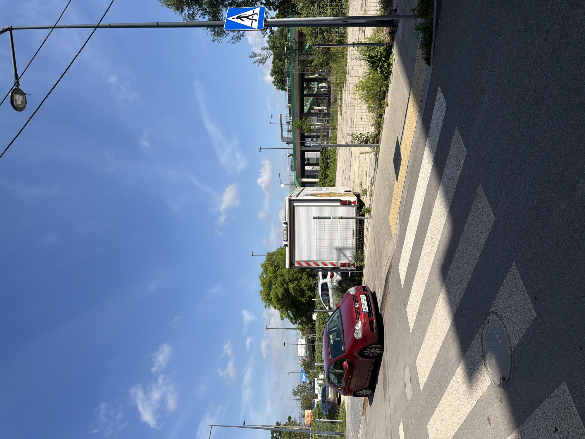 A red Volkswagen Golf is parked on the side of a street next to a pedestrian crossing in front of a white delivery truck. The street is paved with asphalt and has white crosswalk markings. The parking area beside the street is covered with gravel and a yellow tactile paving strip is visible. In the background, there is a multi-level overpass with greenery and a few cars and pedestrians. A blue pedestrian crossing sign is mounted on a pole on the right side of the image. The sky is blue with scattered white clouds.