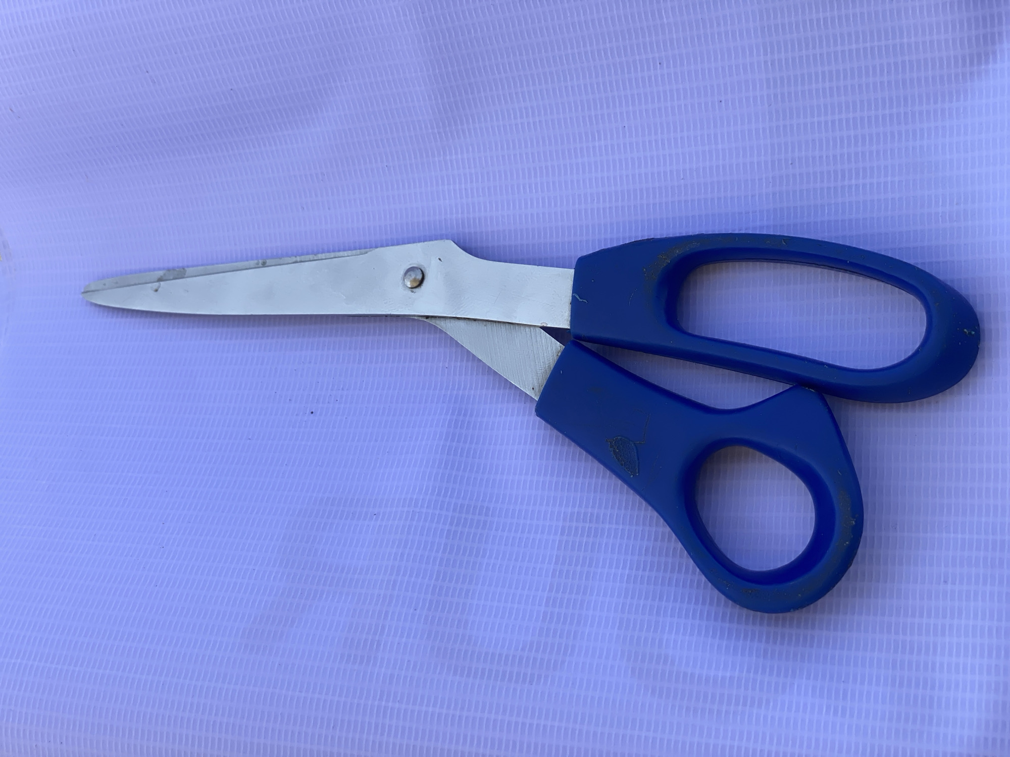 A pair of blue-handled scissors lies open on a light purple, textured surface. The blades of the scissors are silver and appear to be made of metal. The right handle is larger and more elongated, designed to fit multiple fingers, while the left handle is smaller, intended for the thumb. The surface beneath the scissors has a woven pattern, with thin vertical lines creating a grid-like effect. The lighting suggests it is daytime, possibly outdoors, given the overall brightness of the image. There is no visible text in the image. The scene is simple and static, focusing solely on the object and its immediate surroundings.