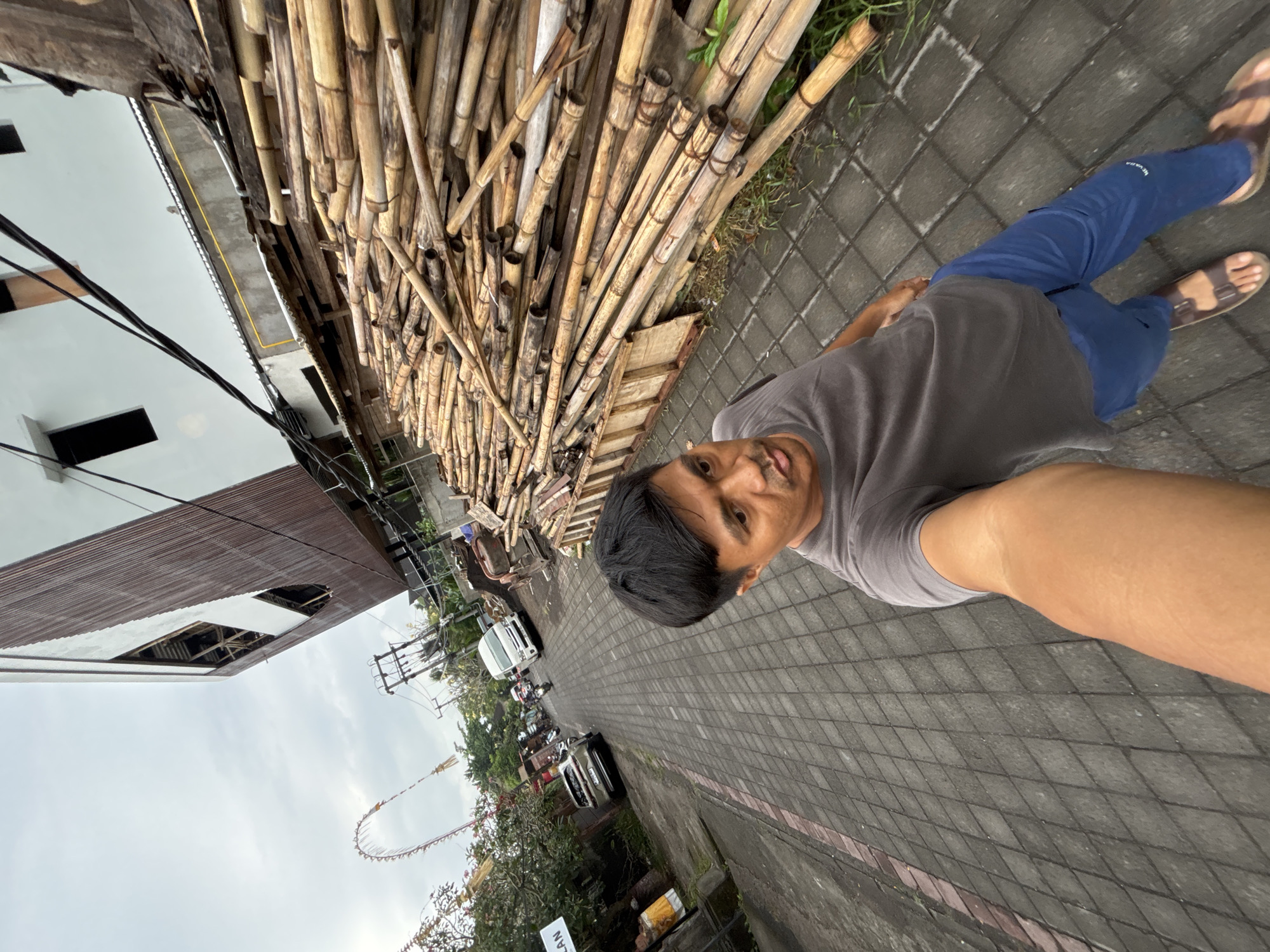 A man is taking a selfie on a street in Ubud District, Indonesia. He is wearing a grey t-shirt, blue pants, and sandals. The street is paved with grey interlocking pavers and has a red brick border on the left. To the right of the street, there is a large pile of bamboo poles. Behind the bamboo is a building with a brown corrugated metal facade and white sections, with dark rectangular windows. In the background, there are more buildings, power lines, a white van, motorcycles, and some greenery. A large, arched bamboo structure is visible in the distance on the left. The sky is overcast.
