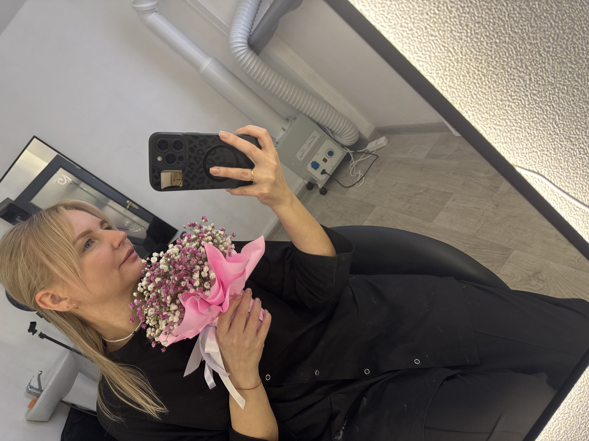 A woman is taking a selfie in a room with a mirror. The woman has blonde hair and is wearing a black robe. She is holding a bouquet of small white and pink flowers wrapped in pink paper and tied with white ribbon. The woman is wearing a ring on her left ring finger. The phone she is using to take the selfie has a patterned case. In the background, there is a white wall with a white ventilation duct and a gray air filtration unit with a power cable. There is a black chair and a section of a black-framed mirror on the left. Based on additional information, the location is Chaikovsky, Russia.