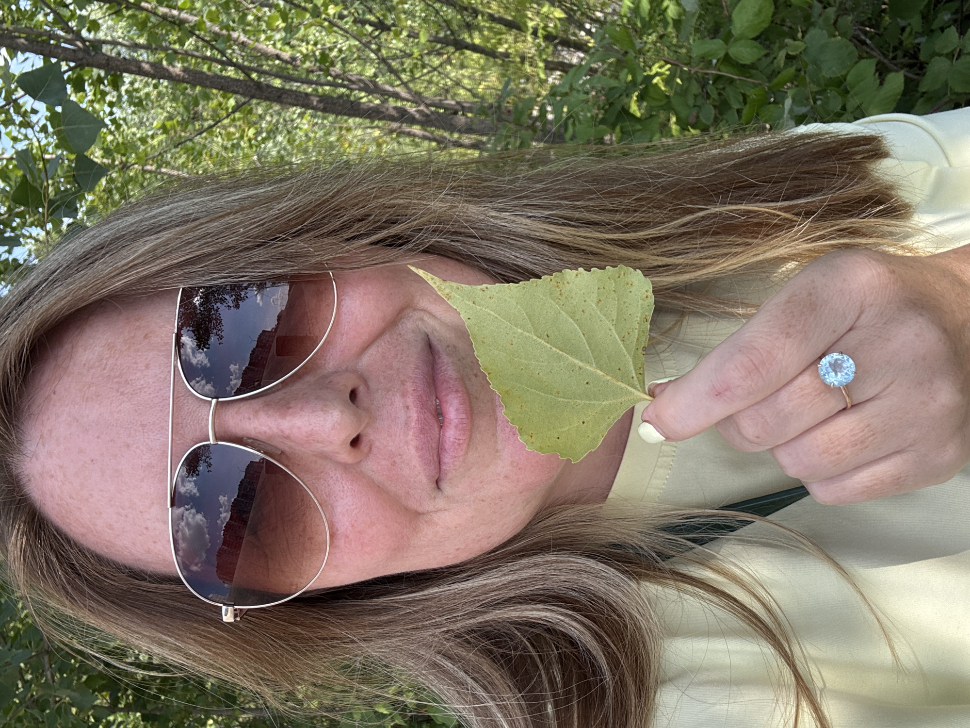 A medium shot shows a woman with blonde hair and fair skin, wearing large aviator sunglasses and a light yellow shirt. She is holding a single green leaf in her right hand, which is adorned with a large, light blue stone ring. The background is filled with lush green foliage and trees, suggesting an outdoor setting, possibly a park or natural area within the city of Ufa, Russia. The lighting indicates it is daytime, with sunlight dappling through the leaves. The woman's face is partially obscured by the leaf and her sunglasses, but her lips are slightly parted in a subtle smile. The image has a relaxed, casual feel.