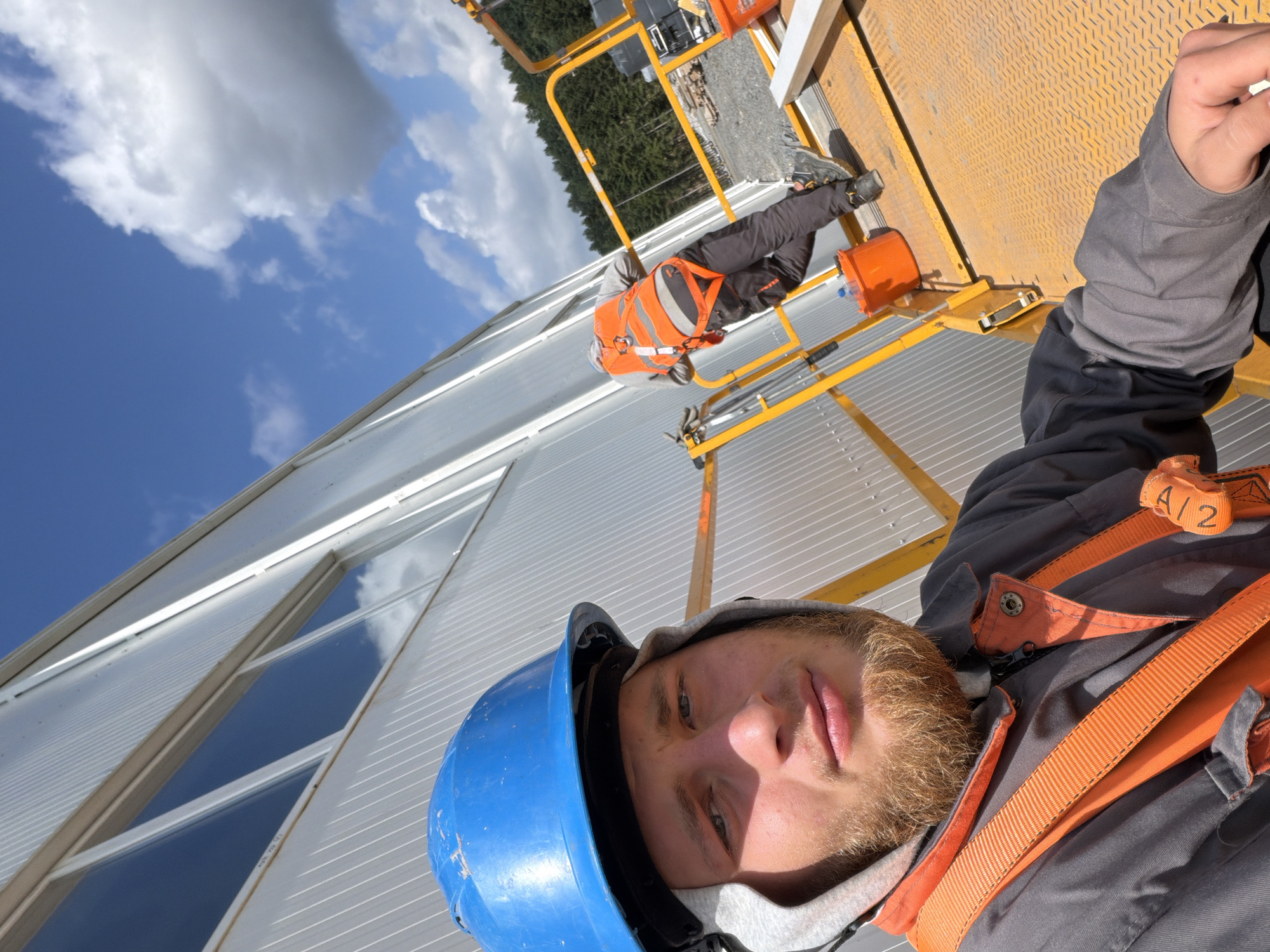 Two construction workers are visible on a yellow elevated work platform. The worker in the foreground is taking a selfie and is wearing a blue hard hat, a grey hooded sweatshirt, a grey jumpsuit, and an orange safety harness. They have a beard and are looking at the camera. The second worker, in the background, is wearing an orange safety vest over a grey sweatshirt, black pants, and dark work boots. They are facing away from the camera and appear to be working on the side of a building. The building has a corrugated metal exterior and several windows. The environment appears to be a construction site, with trees and a forest visible in the distance. The sky is blue with scattered white clouds. The image was taken in Stróża, Poland.