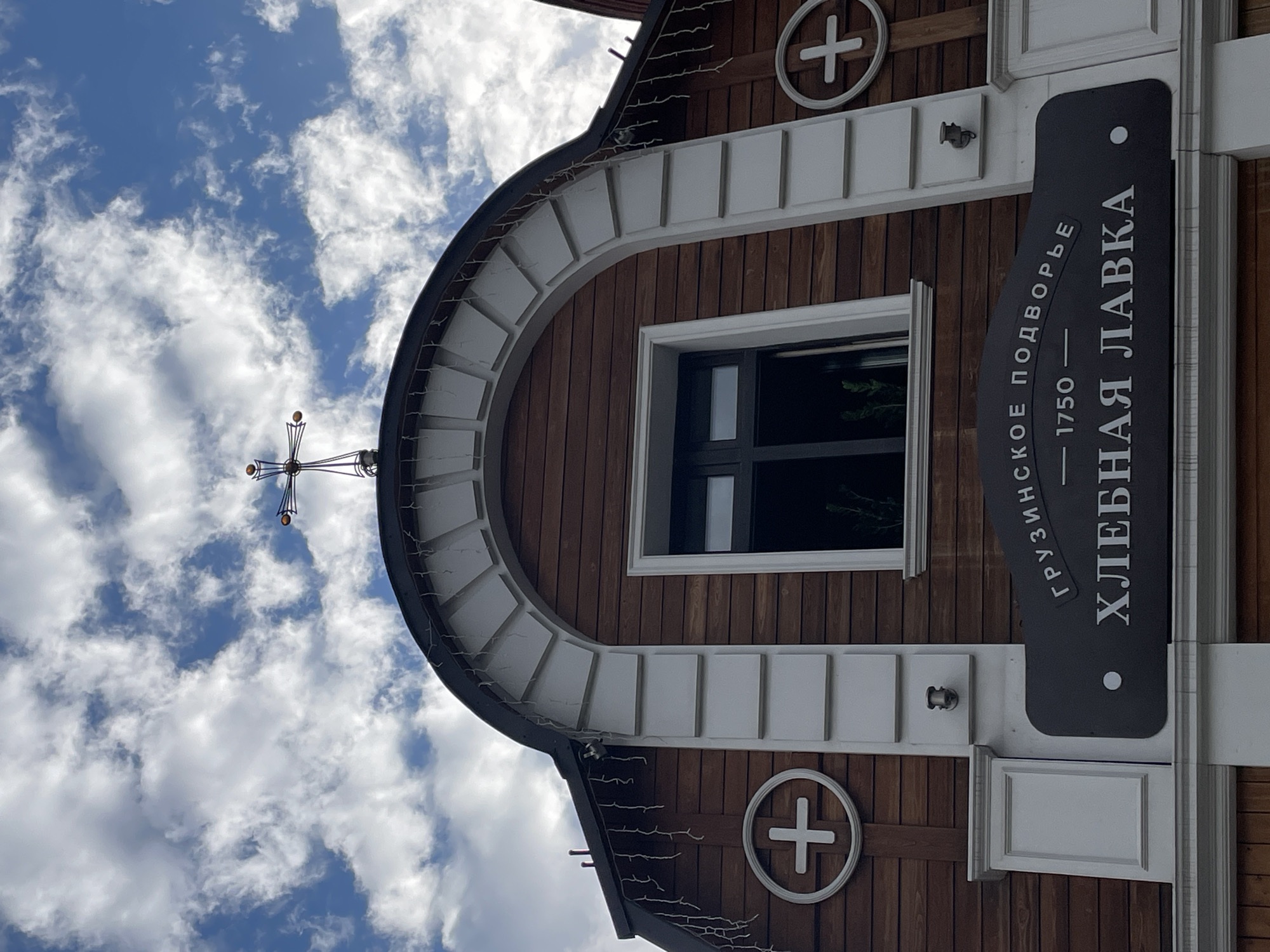 The image shows the facade of a building with a dark brown wooden exterior and white architectural details. A large arched window with dark frames is centrally located on the upper portion of the building. Above the window, a cross is visible against a cloudy blue sky. On either side of the building's upper section, there are circular emblems with a white cross inside. Below the window, a dark sign with white Cyrillic text reads "ГРУЗИНСКОЕ ПОДВОРЬЕ 1750 ХЛЕБНАЯ ЛАВКА", indicating "Georgian Courtyard 1750 Bakery". The building appears to be in a town or city, and the overall aesthetic suggests historical or religious significance. The scene is captured from a low angle looking upwards.