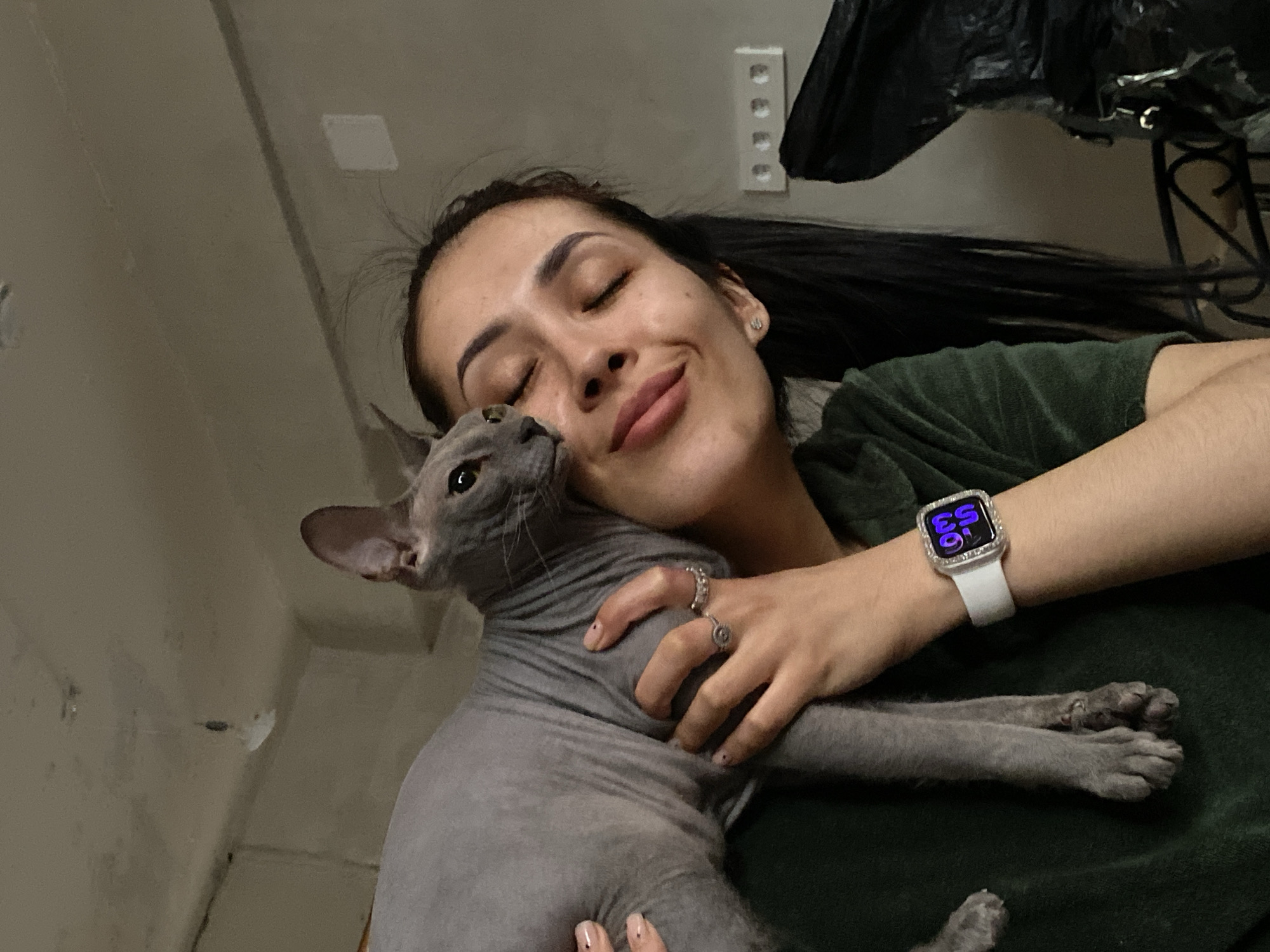 An upward-angled, close-up shot depicts a woman and a Sphynx cat sharing an affectionate moment indoors in Votkinsk, Russia. The woman, with her dark hair pulled back, is seen from the chest up, lying or reclining with her eyes closed and a soft smile on her face, suggesting contentment. She wears a dark green, possibly velvet or fleece, top. Her left arm, adorned with a white smartwatch featuring a sparkly bezel and displaying "03 53" in purple digital numbers, is around the cat. She also has a small stud earring in her left ear and two rings on her left hand, which has light-colored nails with small black dot designs.
The hairless Sphynx cat, with greyish-blue wrinkled skin, is nestled against the woman's neck and chest, appearing comfortable and secure. Its head is turned towards the woman's face, with its eyes open.
The background is a light-colored wall. Visible details in the upper right include a white electrical outlet strip with multiple sockets and a dark, crumpled object, possibly a plastic bag or fabric, hanging above a decorative dark metal stand. The lighting appears dim, suggesting an evening or night setting, consistent with the smartwatch display. The overall scene conveys a warm, intimate, and peaceful interaction between the woman and her pet.