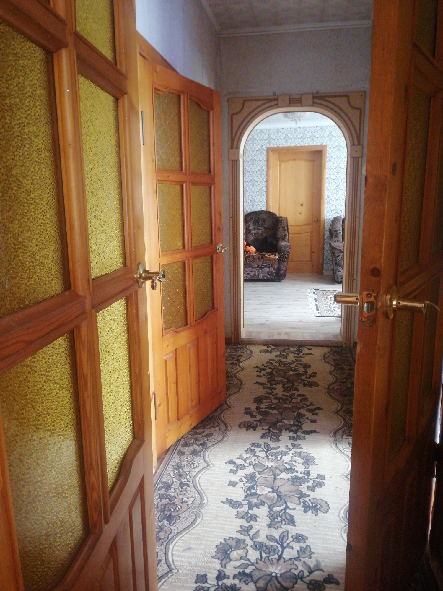 The image depicts an interior hallway of a residential dwelling. The scene is viewed from a low angle, looking down a carpeted hallway that extends into an arched doorway. The hallway is illuminated by overhead lighting, suggesting it is daytime or artificial lighting is used.
The primary subjects are architectural elements and furnishings. Two wooden doors with textured yellow glass panels are prominently featured. One door is open on the left, and another is open on the right. The hallway is adorned with a long, floral-patterned carpet runner. Beyond the arched doorway, another wooden door is visible, as are two upholstered armchairs and what appears to be a small rug on a light-colored floor. The wallpaper in the background has a subtle pattern.
No people are visible in the image, nor is any specific activity taking place. The notable details are the style of the doors, the patterned carpet, and the archway, all suggesting a traditional or older interior design. There is no visible text in the image. The overall impression is of a quiet, domestic interior space. The location context provided is Dubrovka, Russia.