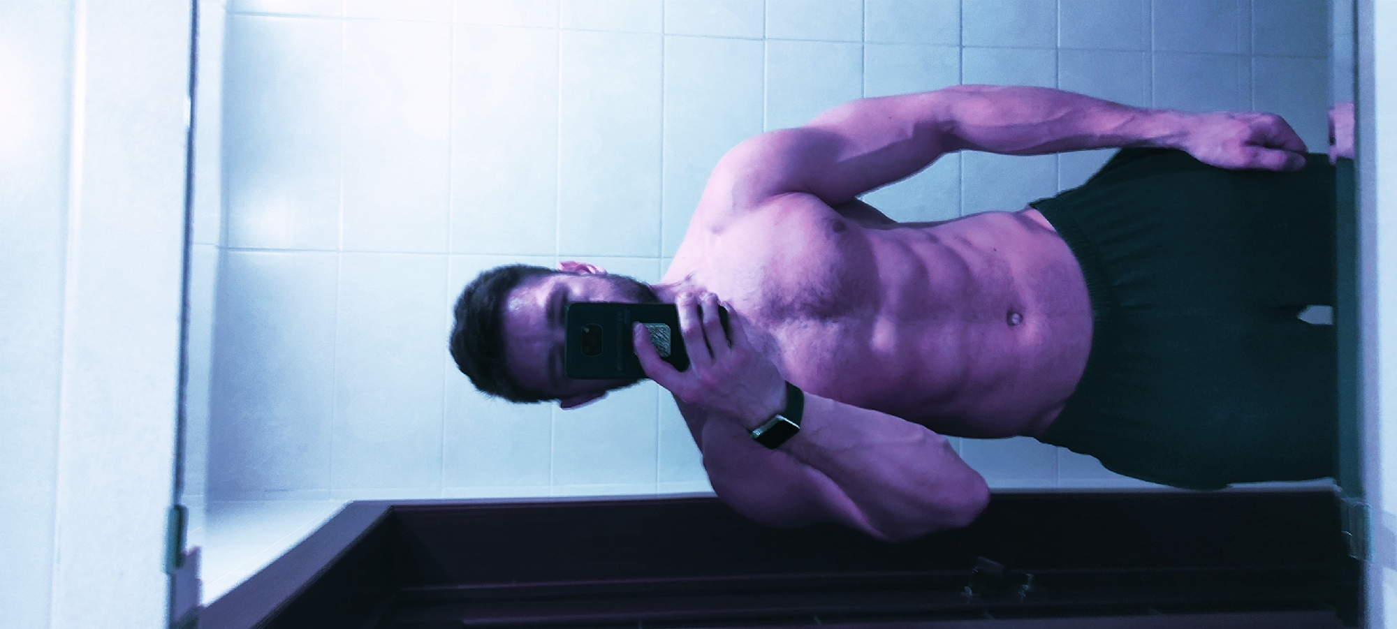 A man takes a mirror selfie in a bathroom. The bathroom has white tiled walls. The man is shirtless, wearing dark shorts, and holding a smartphone. He is wearing a smartwatch on his left wrist. The scene's location cannot be determined as Fryazino, Russia, from the visual information provided.
