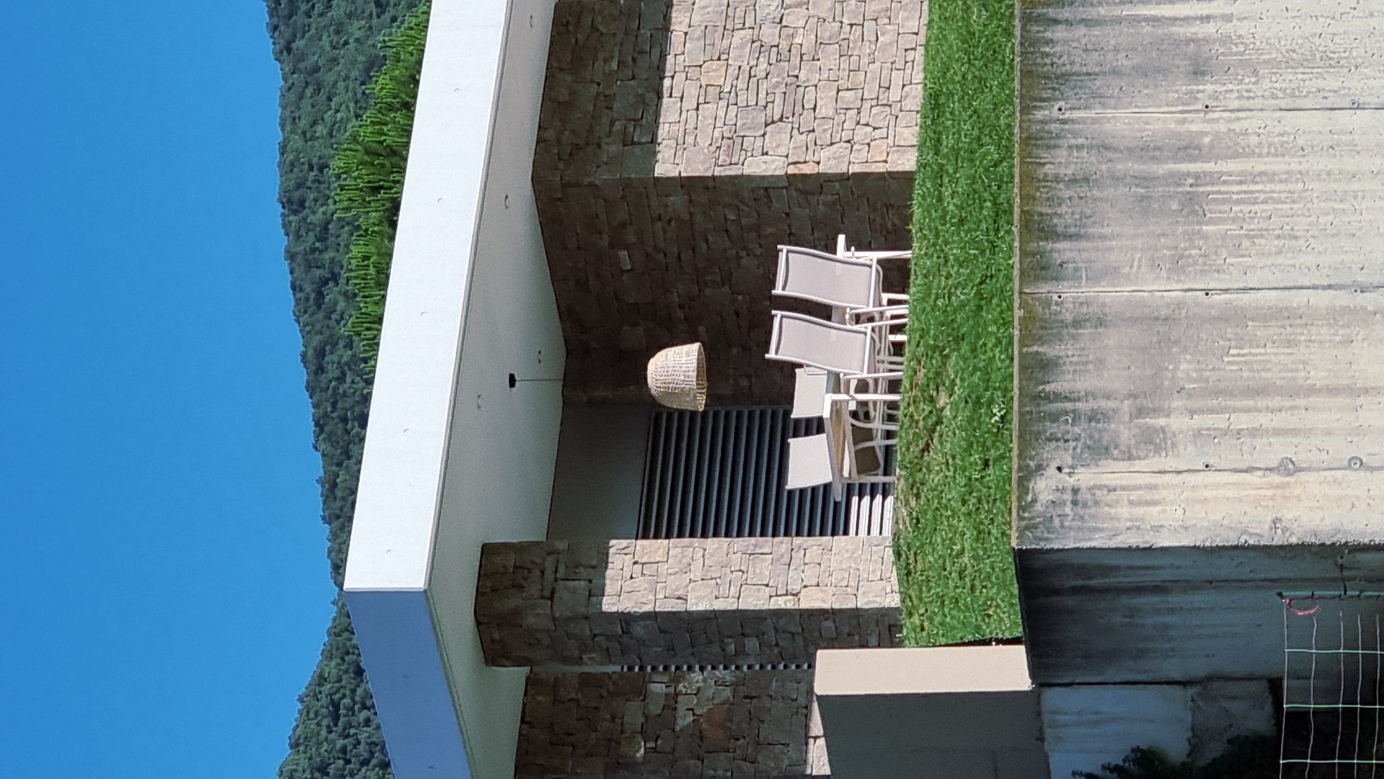 The image captures an outdoor seating area with a table and four chairs on a grassy slope in front of a modern stone building. The building features a white overhang supported by stone pillars. A woven pendant light hangs above the table. In the background, lush green mountains rise against a clear blue sky. The scene appears to be a tranquil setting, likely in Sant Joan les Fonts, Spain, given the architectural style and natural landscape.