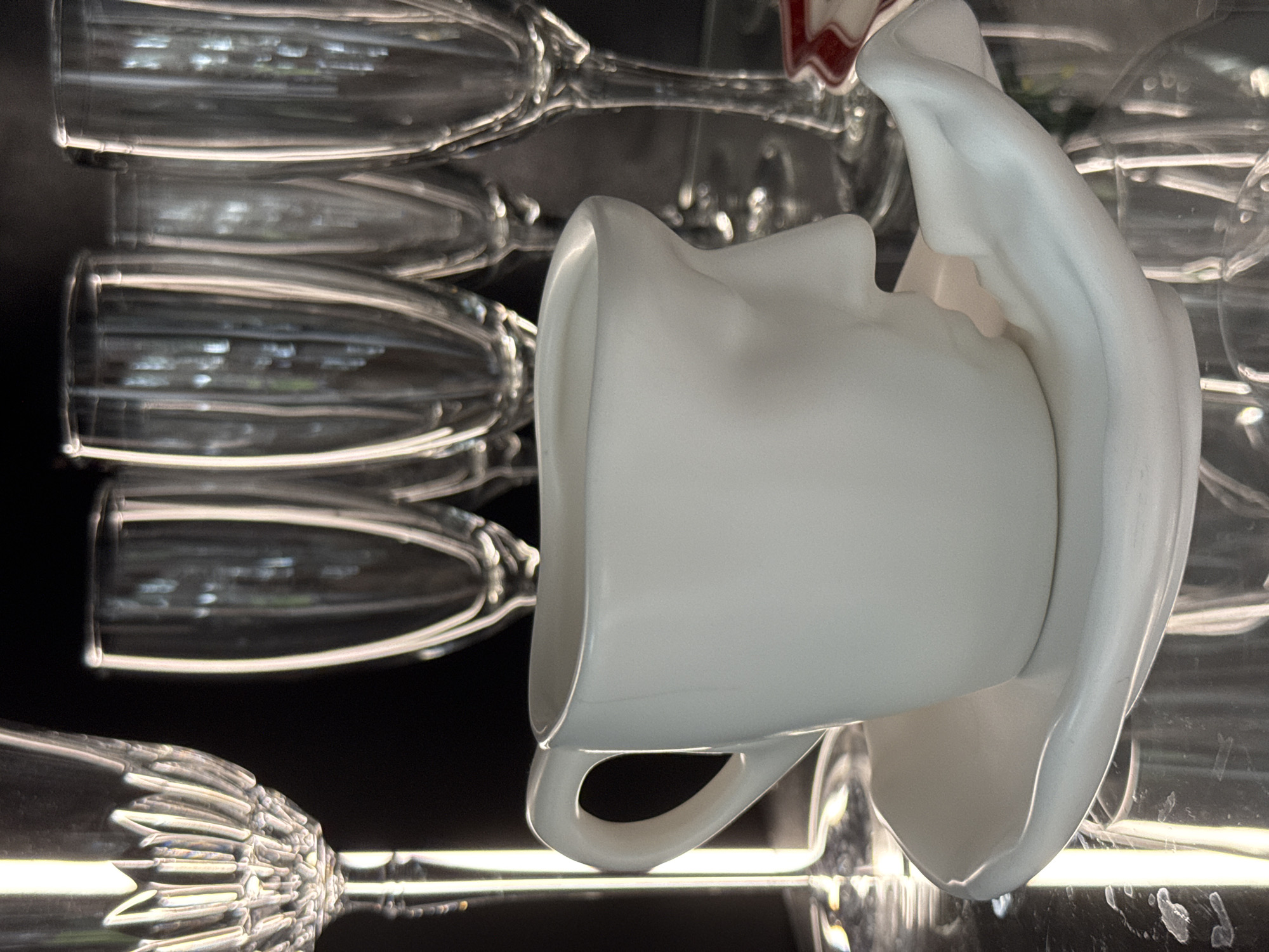 This media file features a close-up, high-angle shot of a white ceramic mug with a unique, sculpted design that resembles a face, placed amongst several clear stemmed glasses. The mug has a large handle and its body is contorted, creating a sculptural effect. The surrounding glasses are stacked and appear to be of various shapes, with some having fluted bases and others having smooth bowls. The lighting is dramatic, with strong highlights reflecting off the glassware and the ceramic surface, creating a sense of depth and texture. The background is dark, which further emphasizes the objects in the foreground. The overall impression is one of still life arrangement, possibly showcasing decorative tableware. There are no people visible, and the scene appears to be indoors, likely in a studio or a setting where glassware is displayed. The image's orientation is unusual, with the subjects appearing sideways. No text is visible in the image. The scene's context in Steklyannyi, Russia, doesn't offer specific visual cues within the image itself, but suggests the origin of the image.