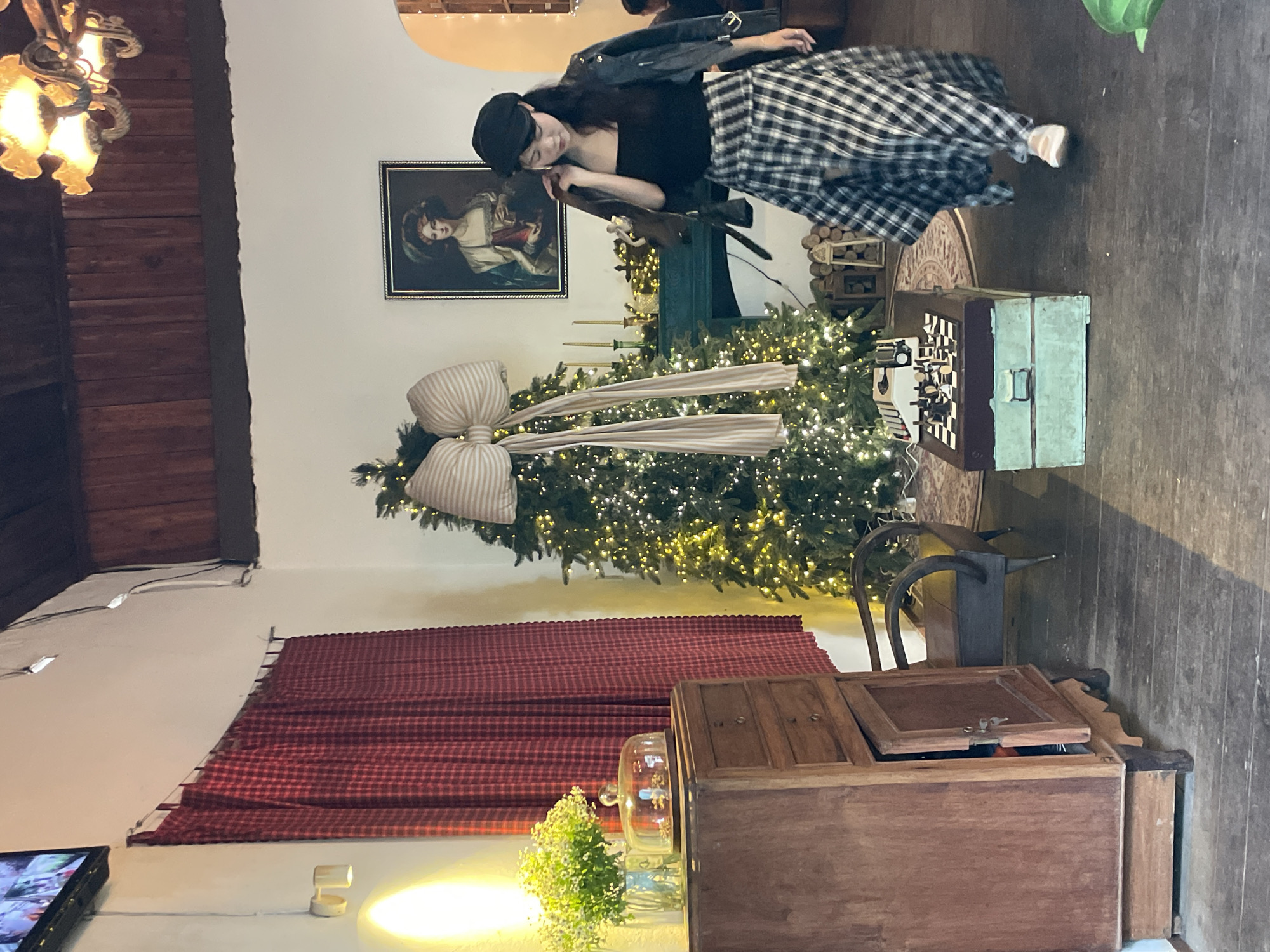 The image depicts an indoor scene, likely a cafe or a room decorated for the holidays in Da Lat, Vietnam. A woman, dressed in a black strapless top, a leather jacket draped over her shoulders, a black cap, and a black and white plaid skirt, is walking from right to left across the frame. She is wearing light-colored slip-on shoes.
In the background, a decorated Christmas tree with numerous small lights and a large striped bow is visible. To the left of the tree is a wooden dresser with several drawers, one of which is open. On top of the dresser, there is a vase of white flowers and a glass cloche covering some items. A red plaid curtain hangs on the wall behind the dresser.
A painting of a woman in a historical costume hangs on the wall above a dark green mantelpiece. On the mantelpiece are several candlesticks. In front of the Christmas tree and to the right of the dresser, there is a small, round rug. On the rug, there is a vintage typewriter placed on top of a light green chest, with a chessboard set up next to it.
The floor is made of dark wooden planks. The overall ambiance suggests a cozy, festive, and somewhat vintage setting. The lighting appears to be a mix of ambient light and the Christmas tree lights, creating a warm glow.