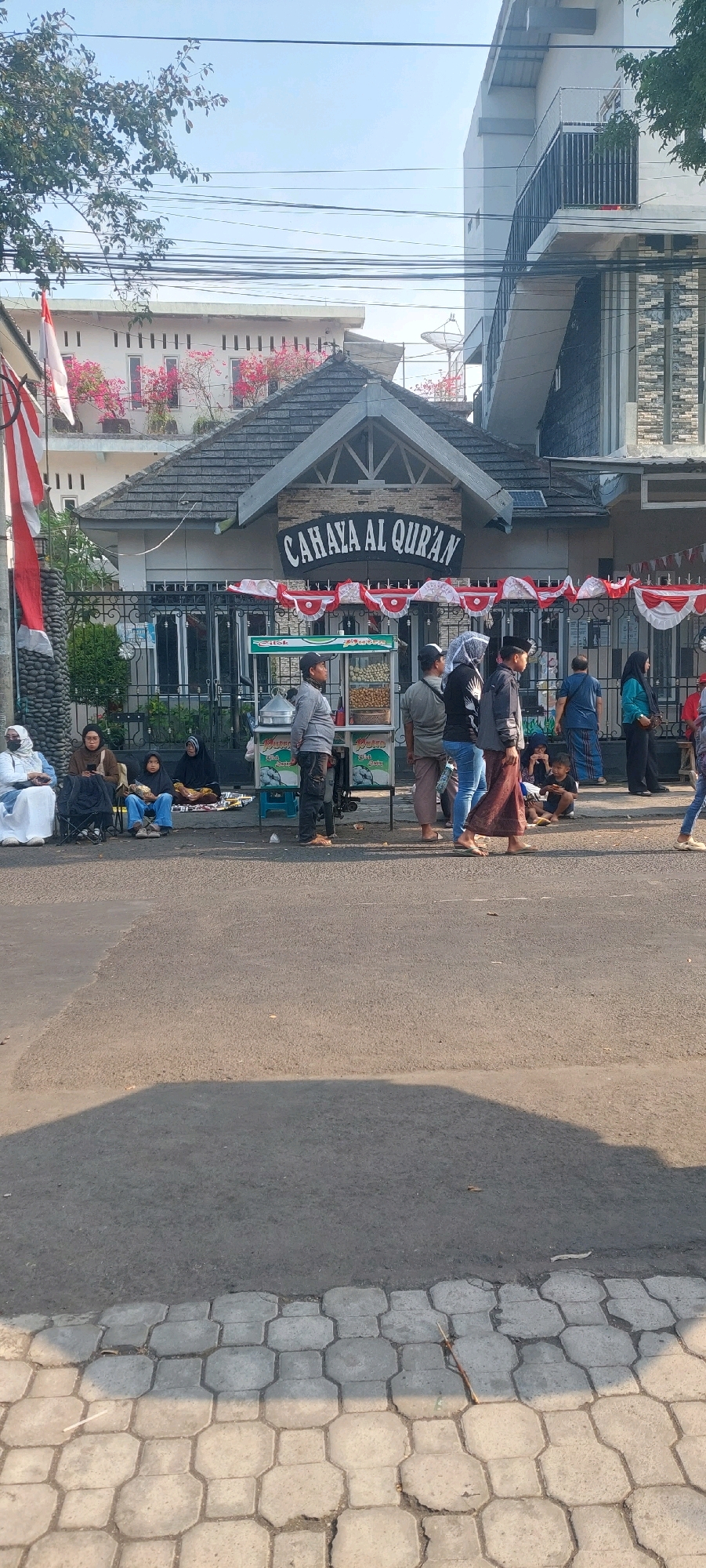 In Jogoyudan, Indonesia, a street scene unfolds in front of a building with the sign "CAHAYA AL QURAN." The building has a gray, textured roof and decorative red and white bunting strung across its facade. In front of the building, a food cart with a green and white canopy displays various fried snacks. Several individuals are present in the scene. On the left, three women are seated on the ground, one wearing a white hijab, another a brown hijab, and a younger girl in a dark hijab. Beside them is a stroller. Closer to the food cart, a man in a gray jacket and dark pants stands facing right. A person in a blue hijab and a man in a gray shirt and brown pants are also near the cart. Further to the right, a woman in a blue hijab and a man in a blue and white patterned garment are standing. A group of children are seated on the ground further down the street. On the far right, a person wearing a blue top and blue jeans is partially visible. The street in the foreground is paved with asphalt, and a textured concrete sidewalk runs along its edge. The lighting suggests it is daytime, with shadows cast on the street.