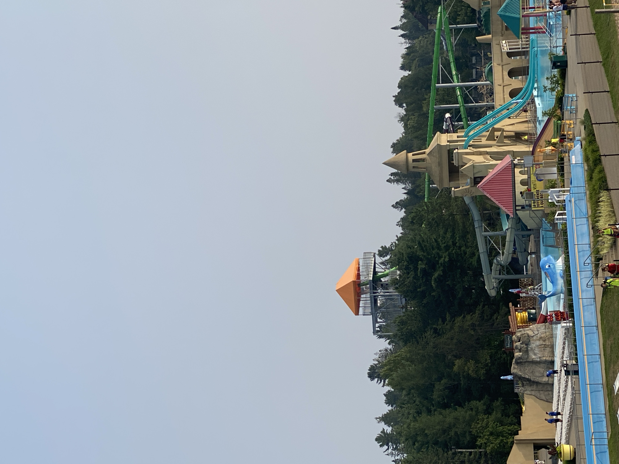 The image shows a wide shot of a water park in Saint-Gabriel-de-Valcartier, Canada. In the foreground, there is a blue pool with a paved walkway around it. Several lounge chairs are visible on the grass area. In the middle ground, a large castle-like structure with turquoise water slides is prominent. A blue inflatable whale floats in a pool adjacent to the castle. To the left, a rock climbing structure with a pool is visible. Behind the water park attractions, a dense forest of green trees forms the background. In the far distance, a tall tower with an orange roof stands above the trees. The sky is clear and pale blue. Several people are scattered throughout the park, some near the pools, others on walkways, and some appear to be staff members in yellow vests. The overall scene depicts a sunny day at a recreational water park.