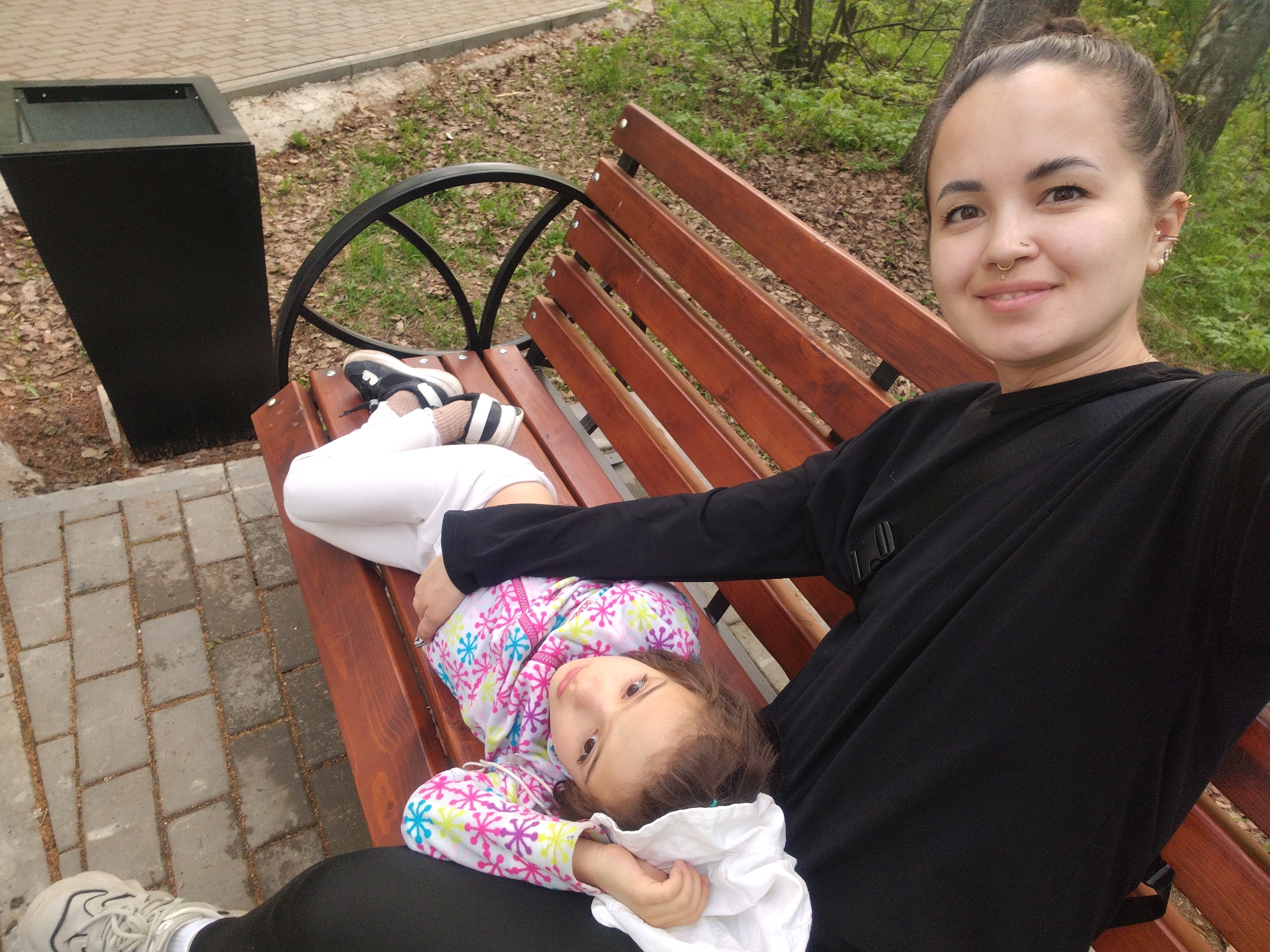 A young woman and a child are sitting on a park bench. The woman is sitting upright, facing the camera, with a slight smile. She has her hair pulled back in a bun, fair skin, and a nose ring. She is wearing a black long-sleeved shirt. The child is lying down on the bench with their head resting on the woman's lap. The child is wearing a colorful patterned jacket. The bench is made of wood slats with a black metal frame. To the left of the bench is a black trash receptacle. The background shows a paved walkway and a grassy area with trees. The location is likely in Noviy, Russia.