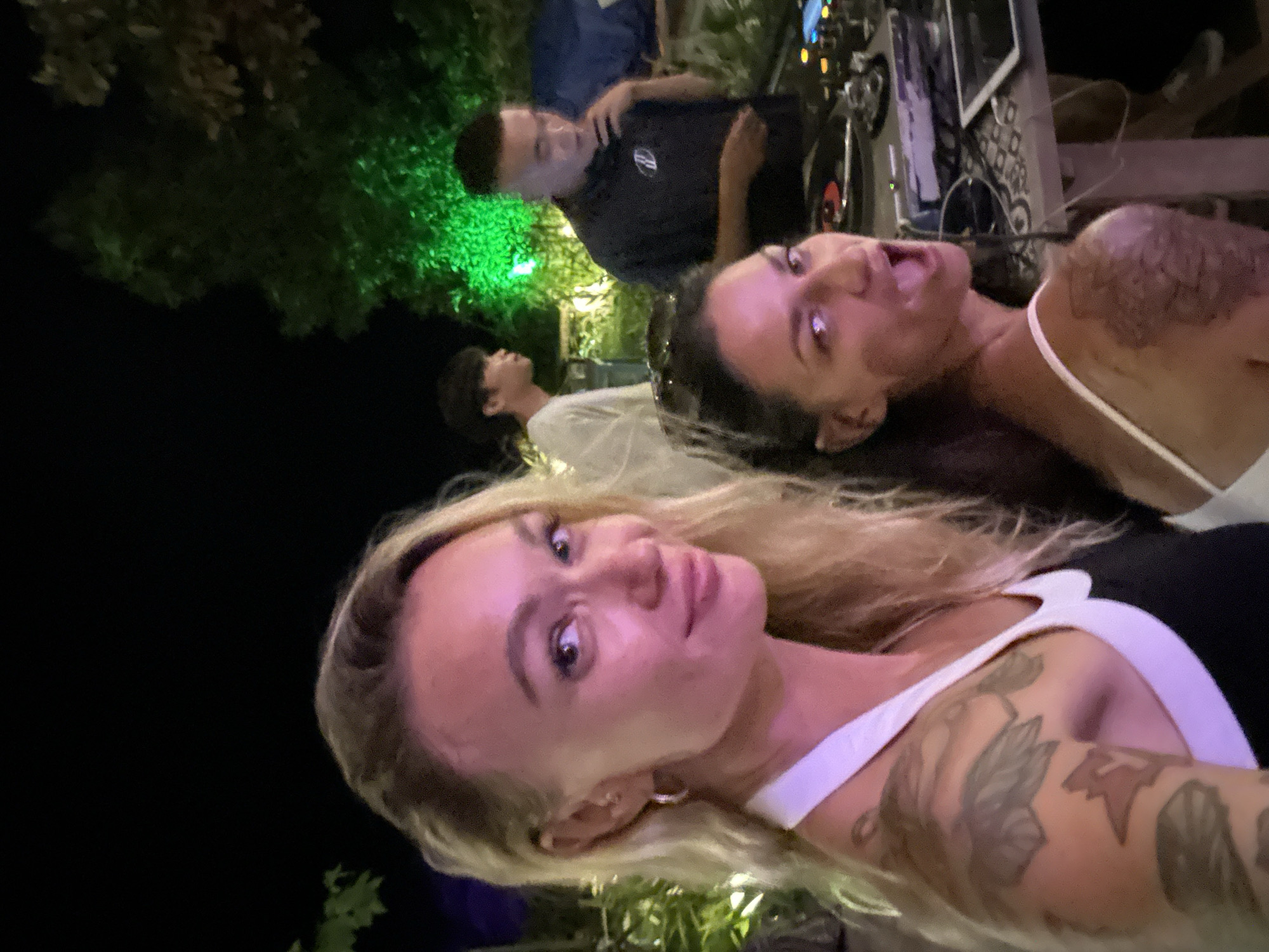 In Mũi Né, Vietnam, at night, two women are in the foreground, positioned at an angle. The woman on the left, with blonde hair, is looking towards the camera with a slight smile. She is wearing a white and black top, with tattoos visible on her arm and shoulder. The woman on the right, with dark hair, is looking over her shoulder towards the camera, her mouth open in a surprised expression. She is wearing a white top and has tattoos on her shoulder. Behind them, a DJ is visible at a console with turntables and equipment, illuminated by green lights. Another person, likely a guest, stands behind the DJ, looking to the side. The background consists of dark foliage and hints of ambient lighting, suggesting an outdoor event.