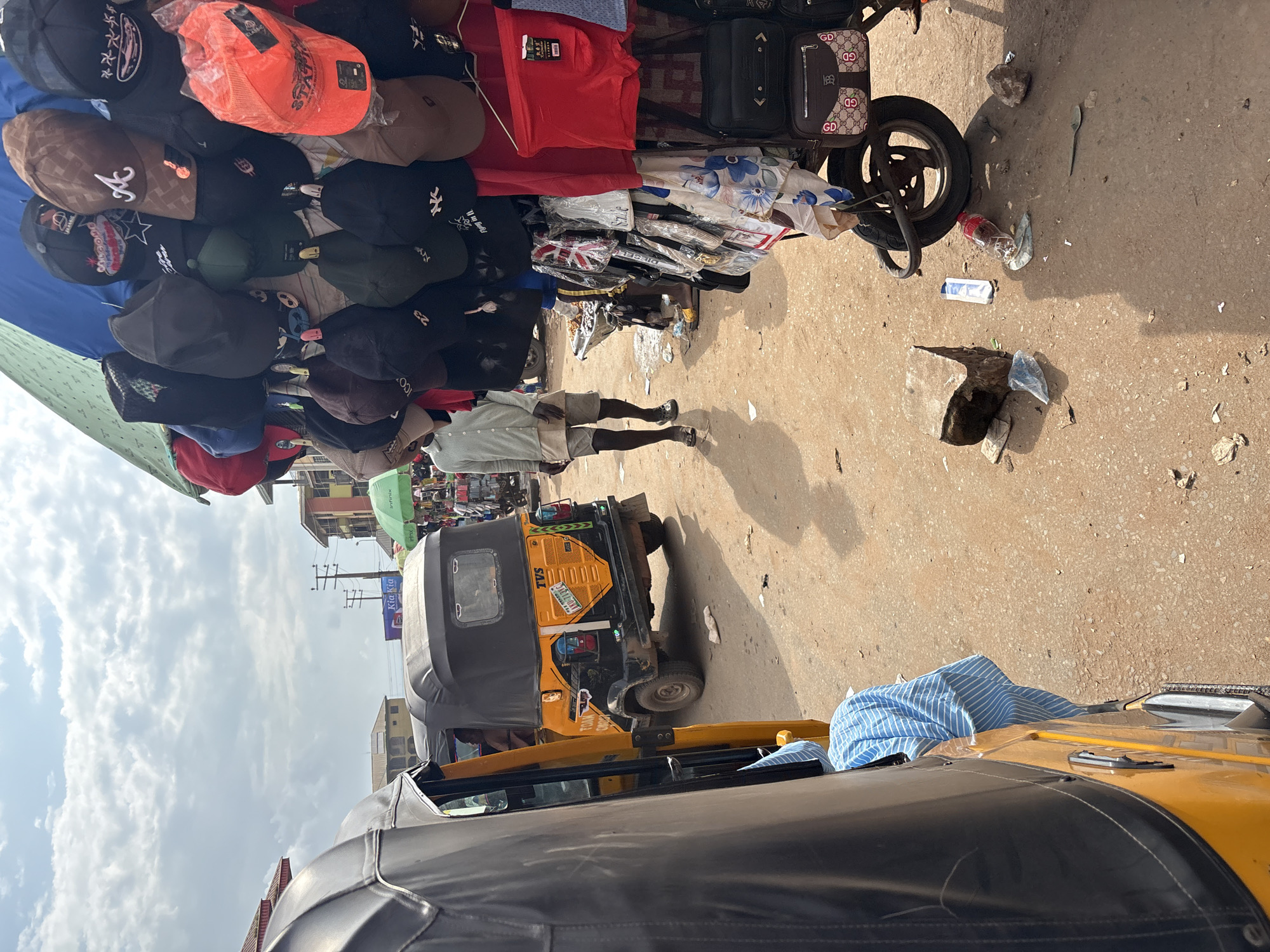 This is a street-level view of a busy market scene in Onitsha, Nigeria. In the foreground, the left side of a yellow and black auto-rickshaw, known locally as a "Keke," is visible. The Keke has a black roof and a blue and white striped cloth draped over its side. To the right of the Keke, an orange auto-rickshaw is parked, also with a black roof. A person is seated in the driver's seat of the orange Keke.
Further into the scene, a market stall displays a variety of goods, primarily caps and bags. Numerous baseball caps in various colors and designs, including brown, black, and orange, are hung from a blue and green tarp. Beneath the caps, several handbags and other accessories are displayed. A man wearing a light green short-sleeved shirt and grey shorts is walking away from the stall, towards the left of the frame, with his back to the camera. He is wearing sandals.
The ground is a dusty, uneven dirt surface scattered with debris, including plastic bags, a discarded water bottle, and rocks. In the background, more market stalls and buildings can be seen, with electrical wires and poles indicating urban infrastructure. The sky above is partly cloudy. The overall impression is of a typical, active market day in a Nigerian city.