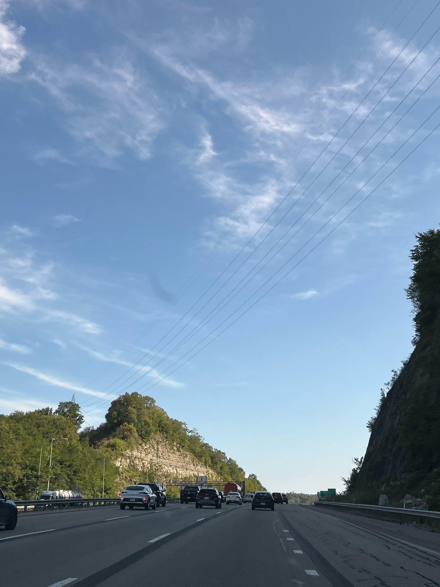 The image captures a view from a vehicle driving on a multi-lane highway in Collier Township, Pennsylvania. The highway has multiple lanes of traffic moving in the same direction. Several cars are visible on the road, including sedans and SUVs. On the left side of the highway, there is a steep, tree-covered embankment with a rocky cliff face. A large truck and a tanker truck are also present in the traffic. On the right side of the highway, another steep, tree-covered hill with a rocky cliff is visible. Overhead, multiple power lines stretch diagonally across the sky, which is a clear blue with some wispy white clouds. The sun appears to be casting shadows, suggesting it is either morning or late afternoon. There are also streetlights and guardrails along the side of the highway. The overall scene depicts a typical highway commute through a scenic, hilly area.