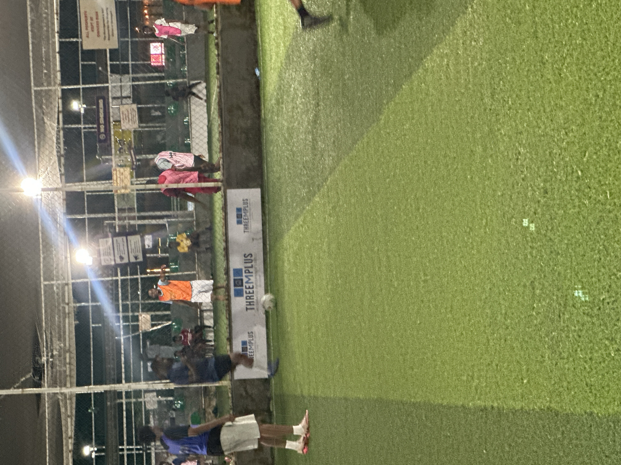 The image depicts an outdoor artificial turf soccer field at night, illuminated by overhead lights. The field is located at the "THREEEMPLUS" sports facility in Abuja, Nigeria. Several individuals are present on or near the field. In the foreground on the left, a person wearing a blue jersey and white shorts stands on the field. To their right, another person in a dark blue jersey and shorts is in motion, possibly kicking or preparing to kick a white soccer ball that is visible on the turf. In the background, several other people are gathered, including individuals wearing orange, pink, and white jerseys. A fence encloses the field, and signs are visible on the fence, including one that reads "NO SMOKING". The overall scene captures a casual soccer game or practice session.