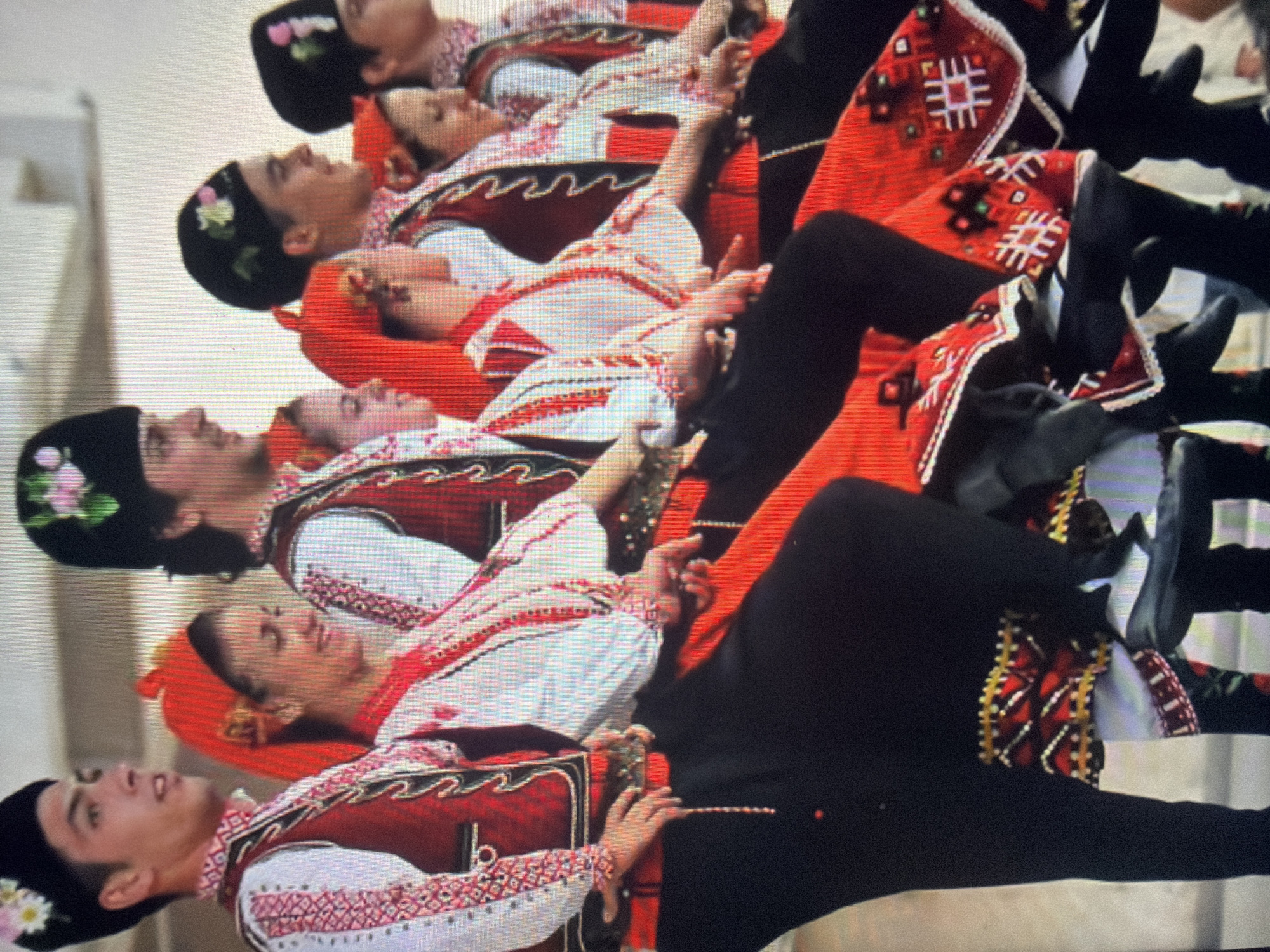This image depicts six young adults, three males and three females, performing a synchronized folk dance in the city of Shumen, Bulgaria. The individuals are arranged in a horizontal line, all facing generally rightward and in similar mid-dance postures with bent knees.
The males wear black, round caps adorned with embroidered pink and white flowers and green leaves. Their attire includes white long-sleeved shirts featuring intricate red and black geometric embroidery on the sleeves and front, over which they wear dark red vests with black trim and gold/white embroidered patterns. Red sashes with metallic elements and hanging chains encircle their waists, paired with black trousers and black shoes.
The females wear red head coverings, white long-sleeved shirts with red and black embroidery, and dark red vests matching the males' attire. They don long red skirts that are heavily embellished with multi-colored geometric patterns (red, white, black, green, gold) around the hem, and black shoes. One female dancer (second from left) displays a smiling expression.
All dancers have their hands positioned near their waists or hips, engaging with elements of their costumes. The performance occurs outdoors under bright, natural light, against a light-colored, out-of-focus background that suggests a simple stage or open area.