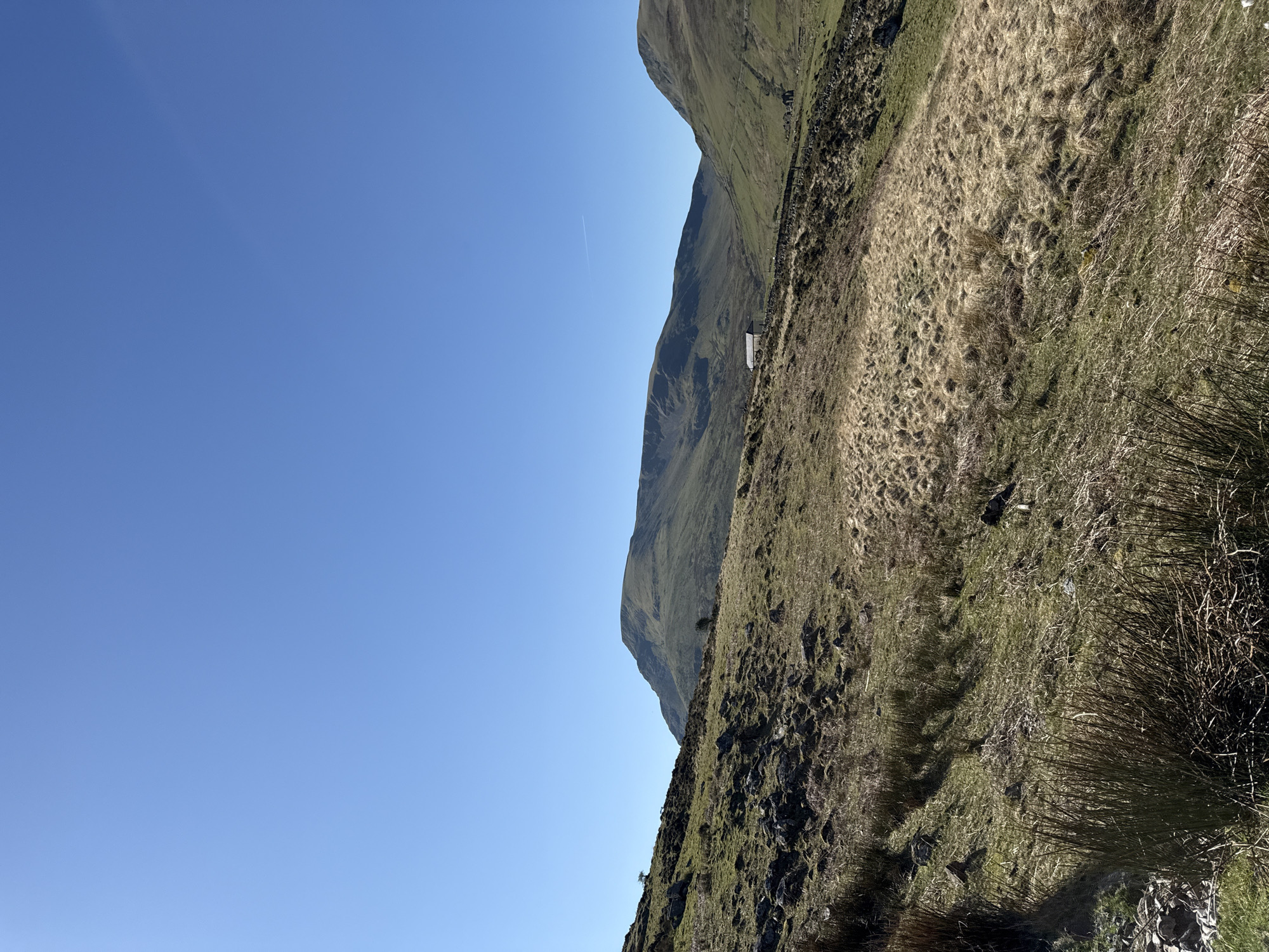 A landscape in Llanberis, United Kingdom features a hillside covered in dry grass and sparse vegetation. A stone wall runs along the slope, and a small, light-colored structure is visible against the hillside. In the background, a steep mountain rises, its face marked with shadows. The sky above is a clear, light blue with a faint contrail. The foreground shows rocky terrain, and the camera angle is somewhat tilted.