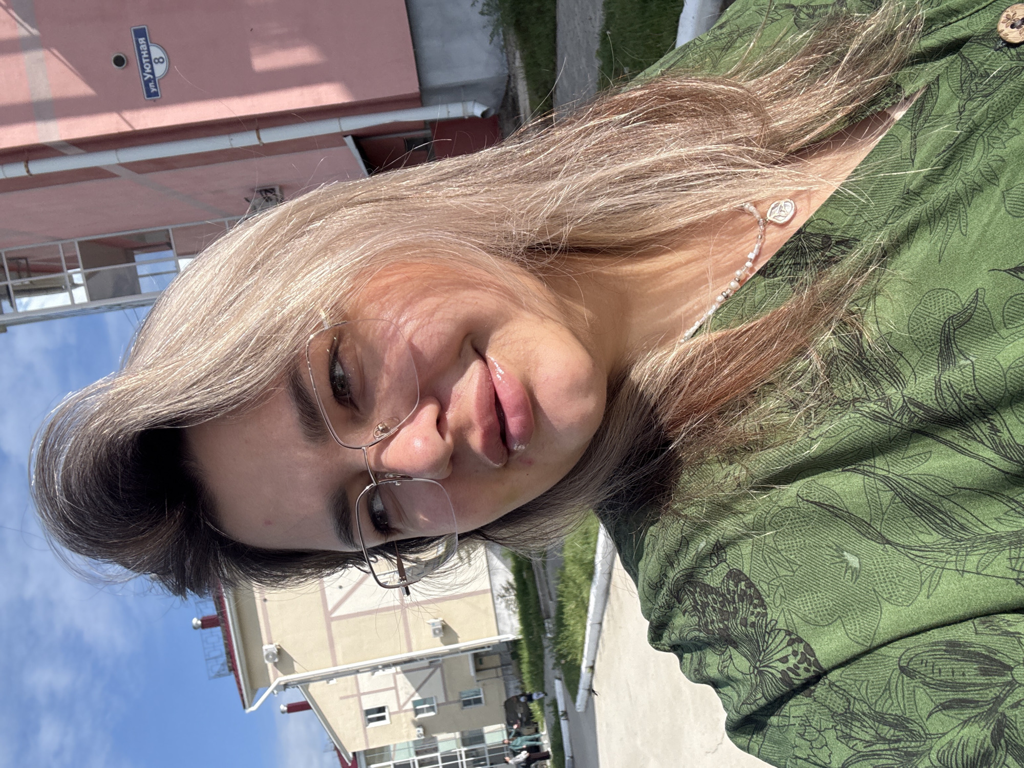A close-up, outdoor, selfie-style photograph shows a young woman with blonde hair and glasses, wearing a green top with a floral pattern and a beaded necklace with a circular pendant. The background features a pink building with a street sign that reads "ул. Уютная 8", suggesting the location is Birobidzhan, Russia. To the left of the pink building is a lighter-colored apartment building. The ground is paved, with patches of grass and some people visible in the distance near the apartment building. The lighting suggests it is daytime, likely sunny, with shadows casting on the woman and the ground.