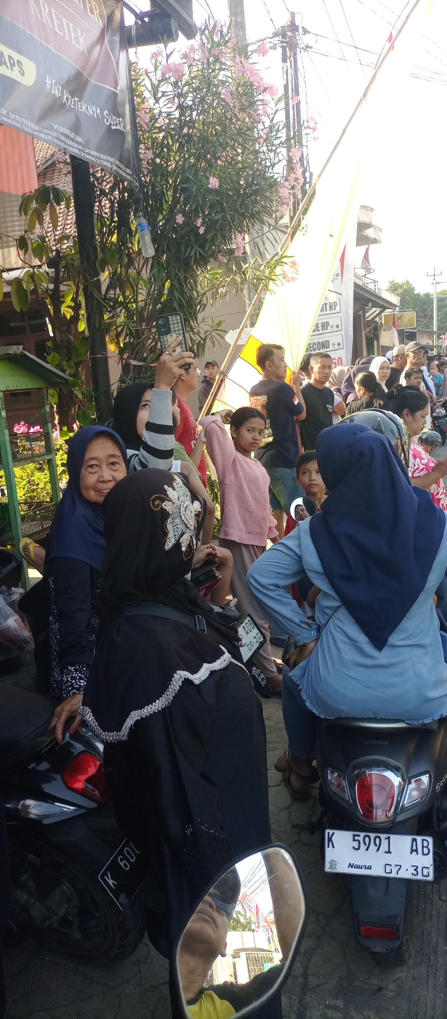 In Jekulo, Indonesia, a group of people are gathered on a street. A woman in a dark blue hijab and a light blue top sits on a dark-colored motorcycle, looking away from the camera. To her left, another woman wearing a dark blue hijab and a black garment with white embroidery is seated on a motorcycle, her face obscured. In the foreground, a motorcycle with a license plate reading "K 5991 AB" is visible. The background shows a crowd of people, some holding a large yellow banner. There are trees and buildings lining the street, and overhead, a network of electrical wires is present. The overall scene appears to be an outdoor gathering or event.