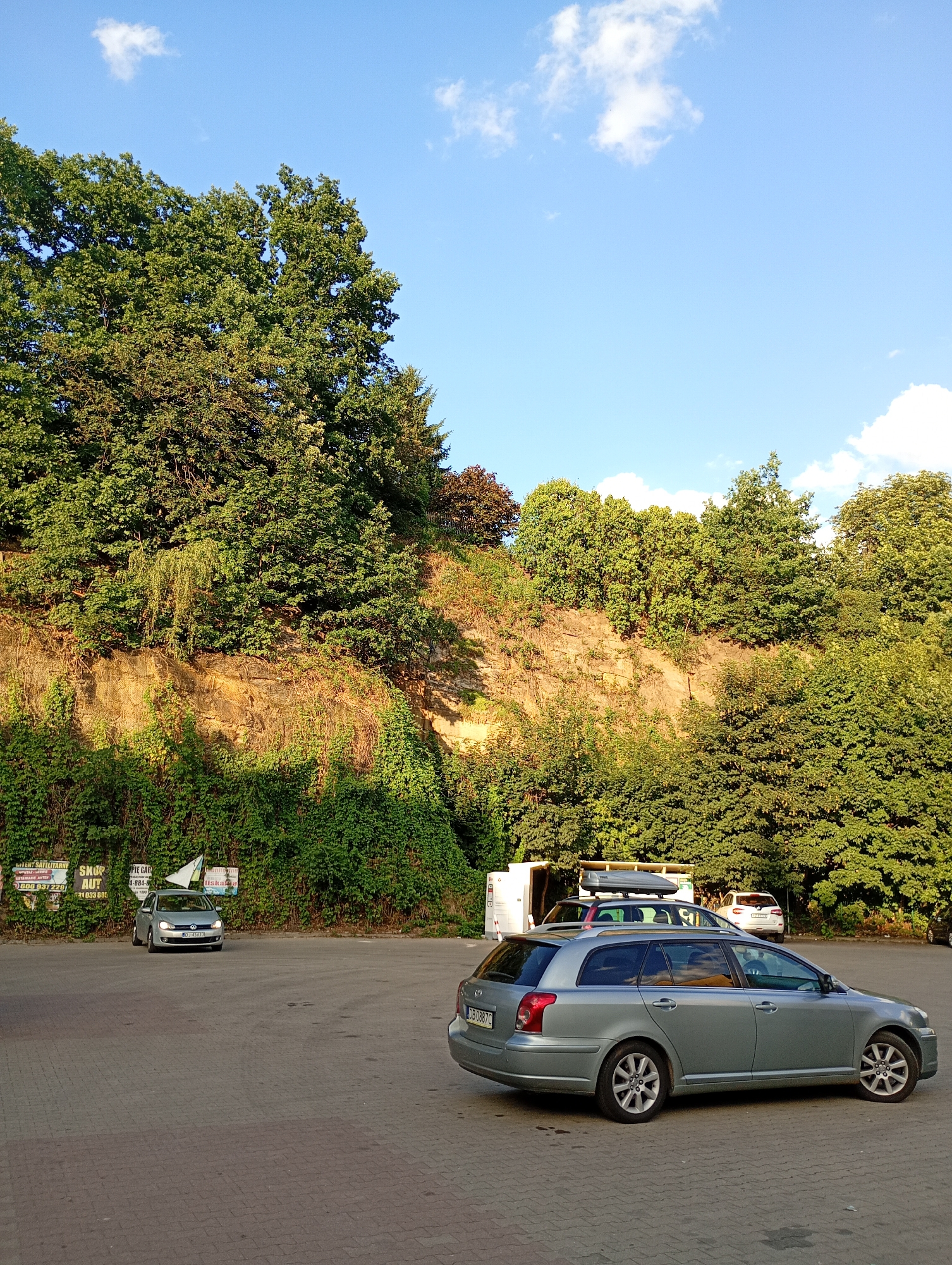 This image depicts a parking lot area adjacent to a vegetated embankment under a clear blue sky with scattered white clouds. The embankment is covered in dense green foliage and appears to be made of rocky, sandy soil. Several cars are parked in the lot, including a silver station wagon in the foreground, a silver hatchback slightly further back with a white flag on its roof, and a white SUV with a roof-mounted cargo box. A white utility trailer is also visible. In the background, a variety of trees form a lush green canopy. Signs are affixed to the embankment in the left portion of the frame. The parking lot is paved with interlocking brick pavers. This scene is located in Wałbrzych, Poland.