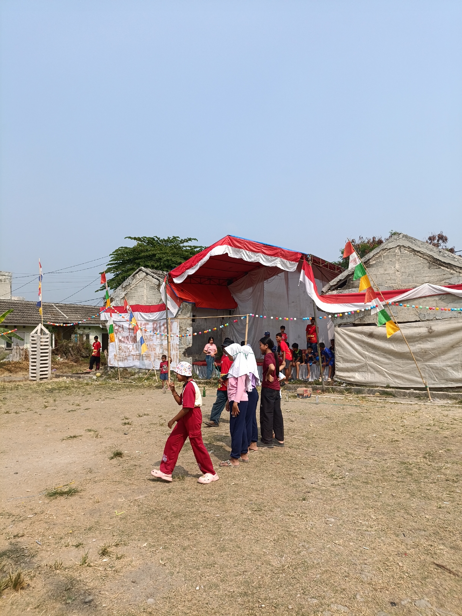 In Cariu, Indonesia, an outdoor event is taking place under a clear sky. A stage draped with red and white fabric serves as the focal point, decorated with colorful bunting. Several people, primarily children and young adults, are present. Some are watching the proceedings, while others are participating in what appears to be a game or performance. In the foreground, a young girl wearing a red outfit and a hat walks across a dusty, grassy field. Several flags, including the Indonesian flag (red and white), are visible, suggesting a celebratory or national occasion. The backdrop includes simple buildings with weathered roofs and some trees.