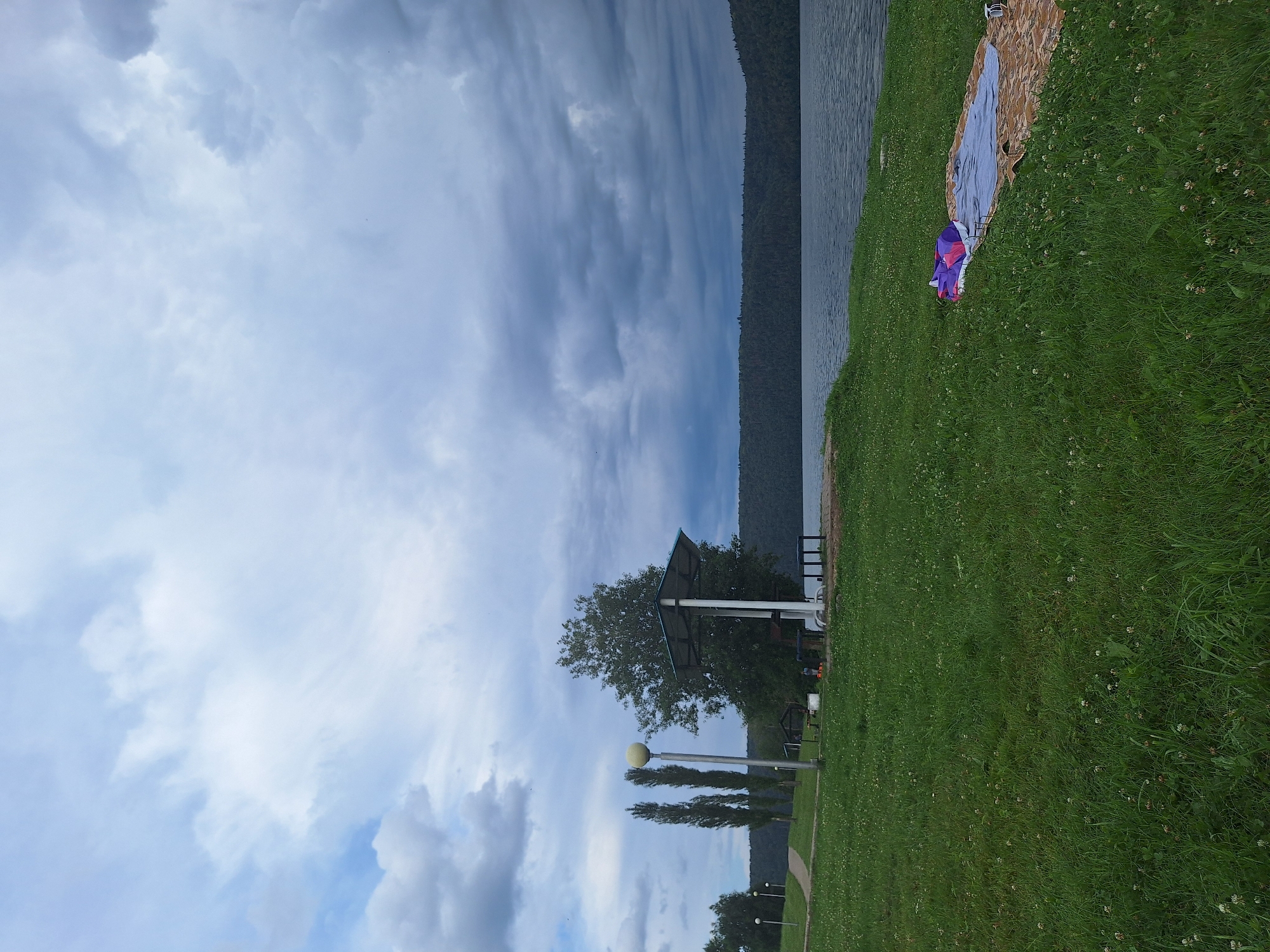 The image depicts a scenic view of a park area near a body of water, possibly a lake or river, in Atamanovka, Russia. The sky is overcast with dramatic, dark clouds, suggesting impending weather. In the foreground, a lush green lawn is dotted with small white flowers. A picnic blanket is spread out on the grass, adorned with a colorful sleeping bag or cushion.
Further back, a paved path winds through the park, flanked by trees and lampposts. A covered pavilion with tables and benches is visible, indicating a recreational area. In the distance, a dense forest lines the opposite bank of the water, creating a natural backdrop. The water itself is calm, reflecting the muted tones of the sky. The overall atmosphere is peaceful and serene, despite the cloudy weather.