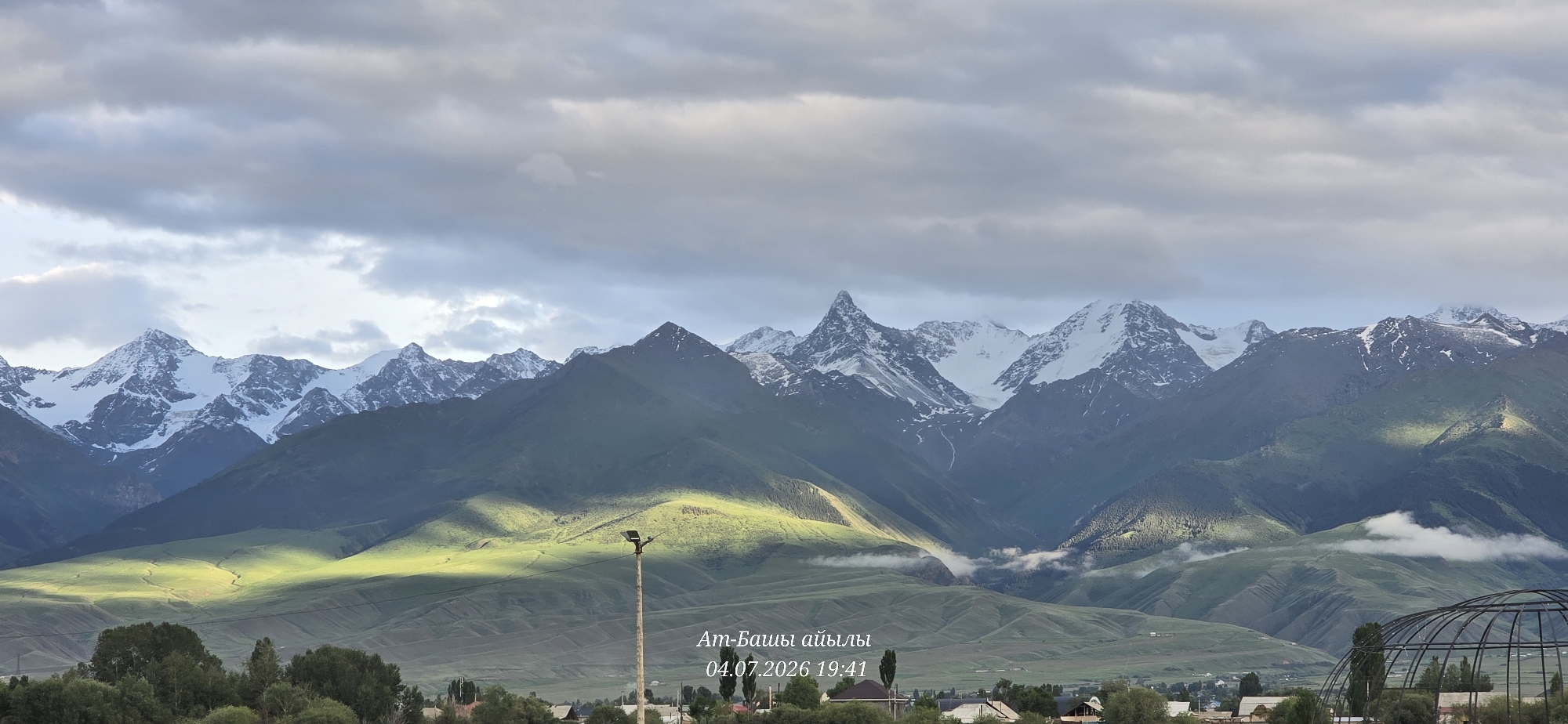 The image depicts a scenic landscape of mountains in At-Bashy, Kyrgyzstan. The sky is partly cloudy, with sunlight casting dramatic shadows across the green, undulating hills in the foreground. Snow-capped mountain peaks rise majestically in the background, their rugged faces partially obscured by wispy clouds and mist. A lone lamppost stands in the middle ground, a solitary man-made structure against the natural grandeur. Below the hills, a village with houses is visible, nestled amongst trees. The timestamp "04.07.2026 19:41" and the text "Ат-Башы айылы" (At-Bashy village) are superimposed on the image, indicating the location and time the photograph was taken.
