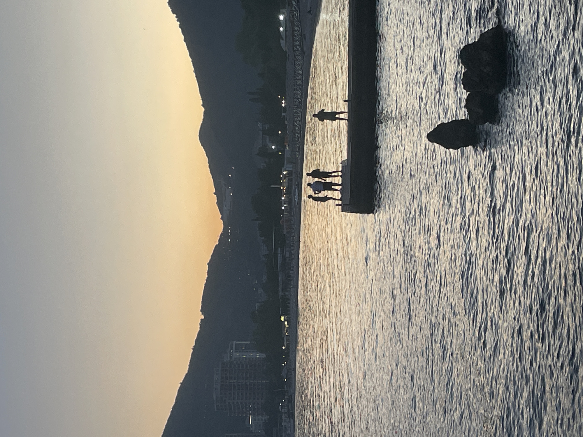 The image shows a waterfront scene in Budva, Montenegro, taken during dusk or dawn. A large mountain range is visible in the background, silhouetted against a colorful sky that ranges from light yellow to pale grey. The shoreline is lined with buildings and streetlights. In the foreground, several people stand on a concrete pier or breakwater that extends into the water. The water surface is textured with ripples and reflections of the light. Further out in the water, there are several large rocks or boulders. The people and objects on the pier and in the water are silhouetted against the lighter water surface. The lighting and composition create a scene of tranquility and natural beauty.