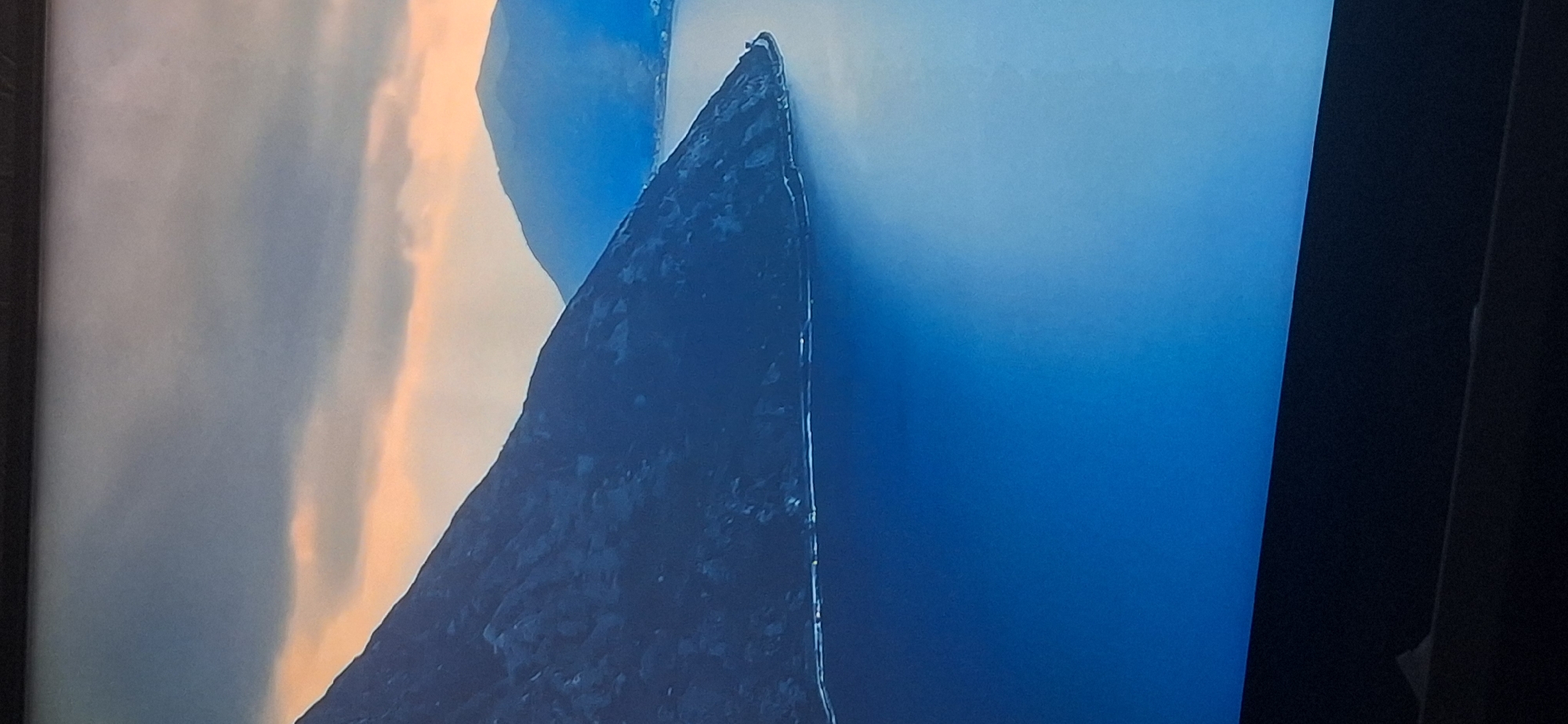 The image depicts a serene natural landscape featuring a large body of water, likely a lake or a fjord, bordered by imposing, dark-colored mountains. The sky above is a dramatic blend of deep grays and oranges, suggesting either a sunrise or sunset. The water is a deep, rich blue, reflecting the colors of the sky and mountains. The mountains themselves are rugged and textured, with visible rock formations. In the distance, smaller, hazier mountains are visible, adding depth to the scene. The overall impression is one of natural grandeur and tranquility. There are no people, objects, or actions identifiable in the image, and the scene is devoid of any urban elements. It is not possible to ascertain the specific location within Bila Tserkva, Ukraine, as the image exclusively portrays a natural, undeveloped environment.