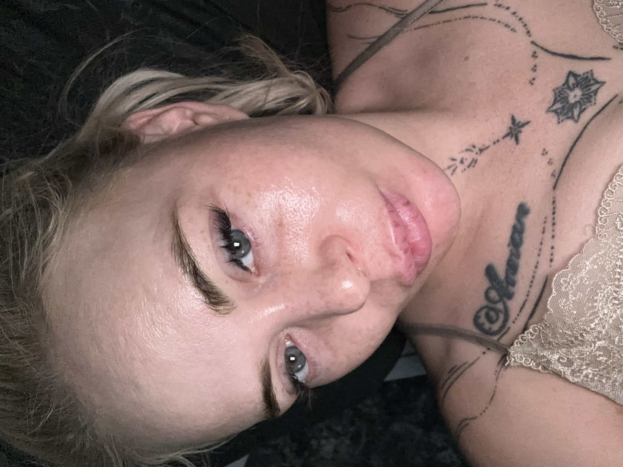 This is a close-up, high-angle, self-portrait of a blonde woman with light blue eyes, light-colored eyebrows, and visible freckles on her face. She is wearing false eyelashes and has full, pink lips. Her skin appears somewhat oily and shiny, with some texture visible on her forehead. She is wearing a light beige lace-trimmed top. There are tattoos visible on her chest and shoulder, with one prominent tattoo on her neck that appears to be text. The background is dark and out of focus. The image appears to have been taken indoors at night.
