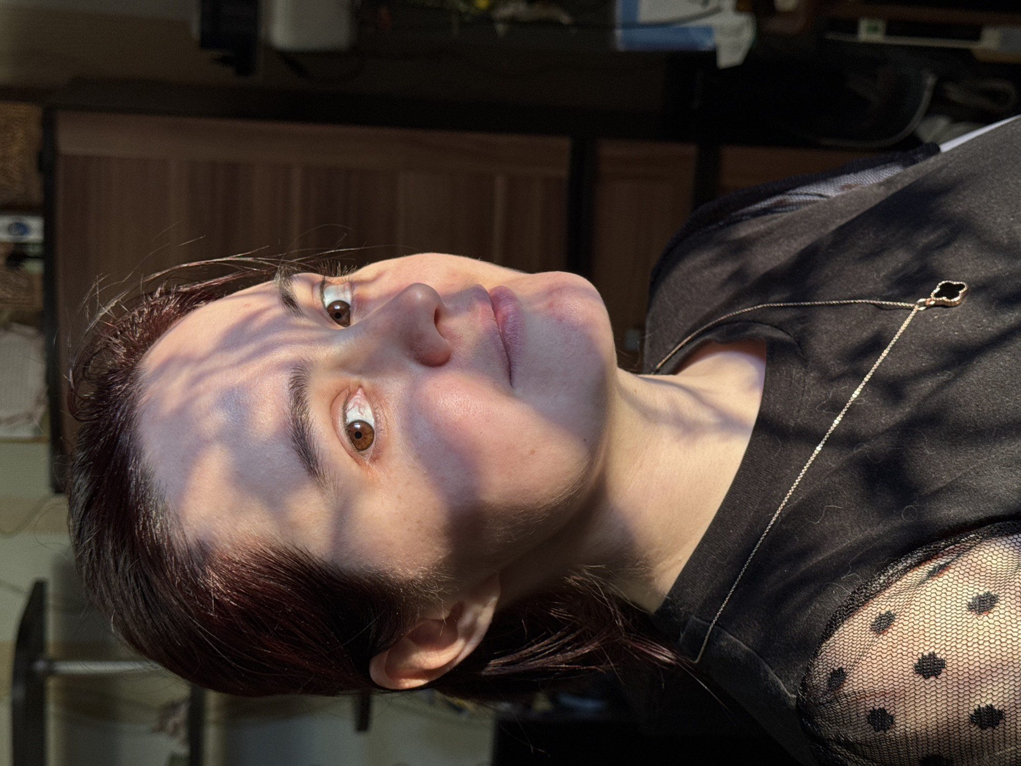 A woman with fair skin and auburn hair is angled upwards in an indoor setting with sunlight creating shadows across her face. She has brown eyes and is wearing a black shirt with a lace sleeve and a gold necklace with a clover pendant. The background is obscured, but shows a wooden panel and various indistinct objects. There is no information to suggest that this image was taken in Izhevsk, Russia.