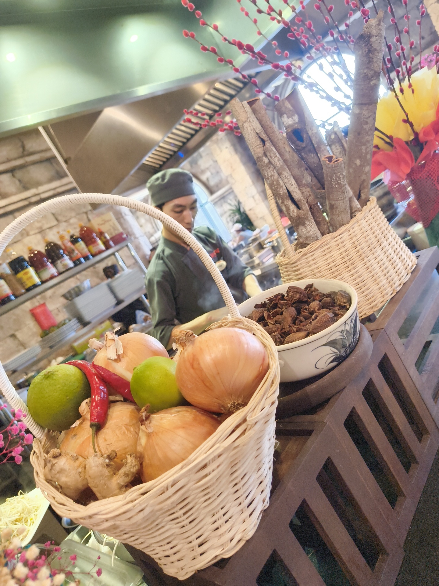 The image shows a basket filled with onions, limes, and chili peppers, with a piece of ginger visible underneath. Behind the basket, a chef in a grey uniform is partially visible, working in what appears to be a kitchen or food preparation area. Shelves with bottles of sauces are on the left, and a bowl of dried spices is on the right, placed next to a basket of branches with red buds. The scene suggests a culinary setting, possibly in Phú Quốc, Vietnam, given the presence of common ingredients and the overall ambiance.