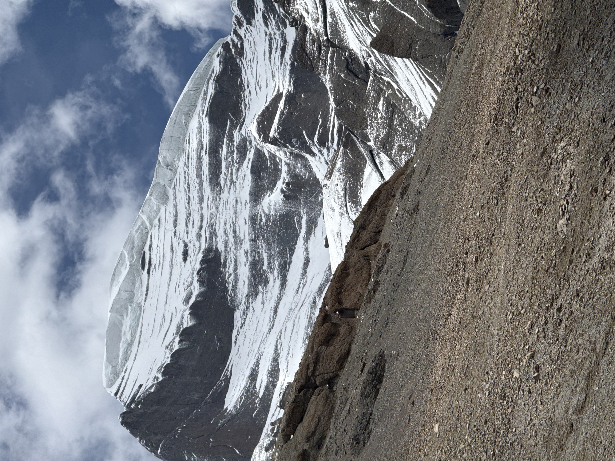 The image shows a steep mountainside with a snow-covered peak against a blue sky with scattered clouds. The left side of the frame is dominated by the snow-covered mountain, with visible crevasses and textured surfaces. The right side is covered in a rocky, gravelly slope. The image is rotated, giving a unique perspective of the mountainous terrain. There is no indication of the location being Parga town, China.