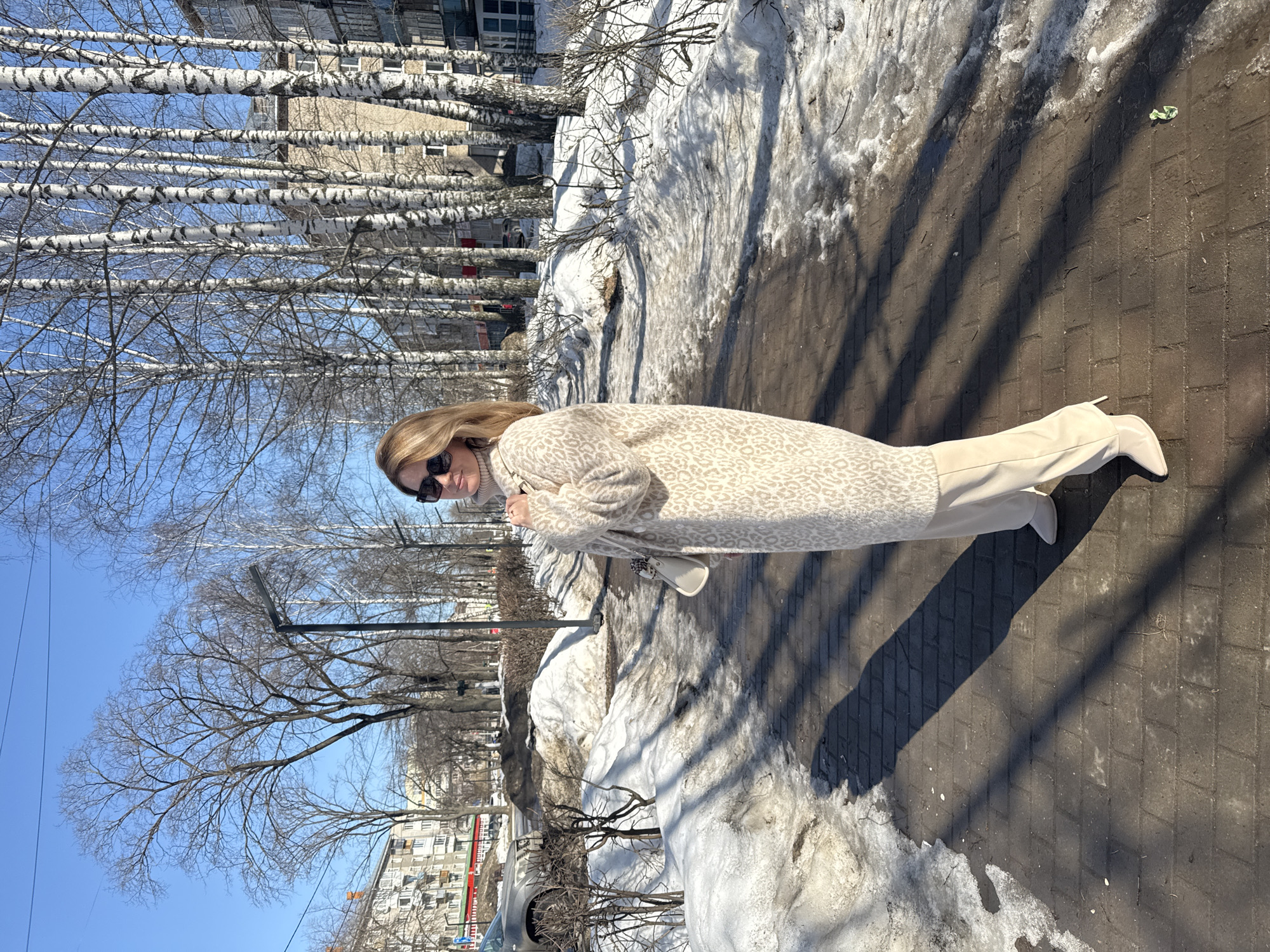 A woman with long blonde hair wearing sunglasses, a patterned beige coat, beige pants, and beige boots stands on a brick sidewalk. Patches of snow are visible next to the sidewalk and near the buildings. The location is in the city of Chaikovsky, Russia, as evidenced by the birch trees and the style of the buildings. The sky is blue and mostly clear. There are bare trees lining the street. In the background, apartment buildings are visible. The sidewalk is paved with gray brick, and the lighting suggests it is a sunny day. The woman appears to be posing for a photograph.