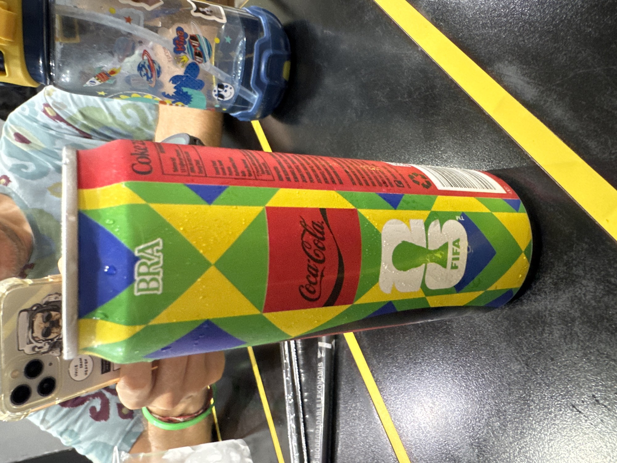 A person is holding a Coca-Cola can with a design featuring the Brazilian flag and FIFA branding, likely as part of a promotional campaign. The person is also holding a smartphone with a phone case showing an image of an astronaut dog. To the left of the can, there is a partially visible clear plastic water bottle with stickers. The background includes a dark surface with yellow stripes, resembling a table or floor. Underneath the can, one can see at least two thin, black pencils. The scene suggests an indoor setting. Without additional information, pinpointing the exact location in Kuta, Indonesia, is not possible.