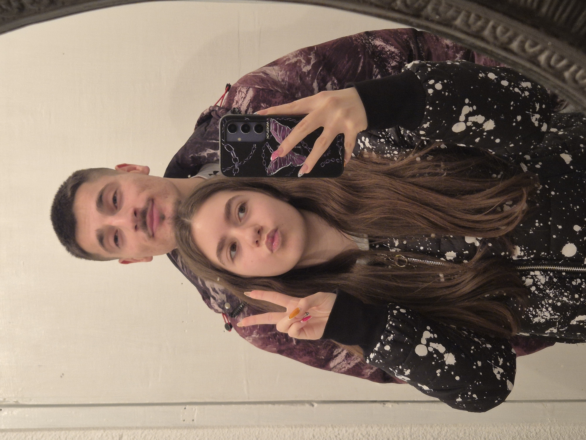 A young man and woman are visible in a mirror selfie. The man has short dark hair and is wearing a purple patterned jacket. The woman has long brown hair and is holding a black phone with a purple butterfly design. She is wearing a black jacket with white splatter marks and is making a peace sign with her hand. The background appears to be a white wall with a ledge. The location is Pavlikeni, Bulgaria, and there are no indications in the image to verify.