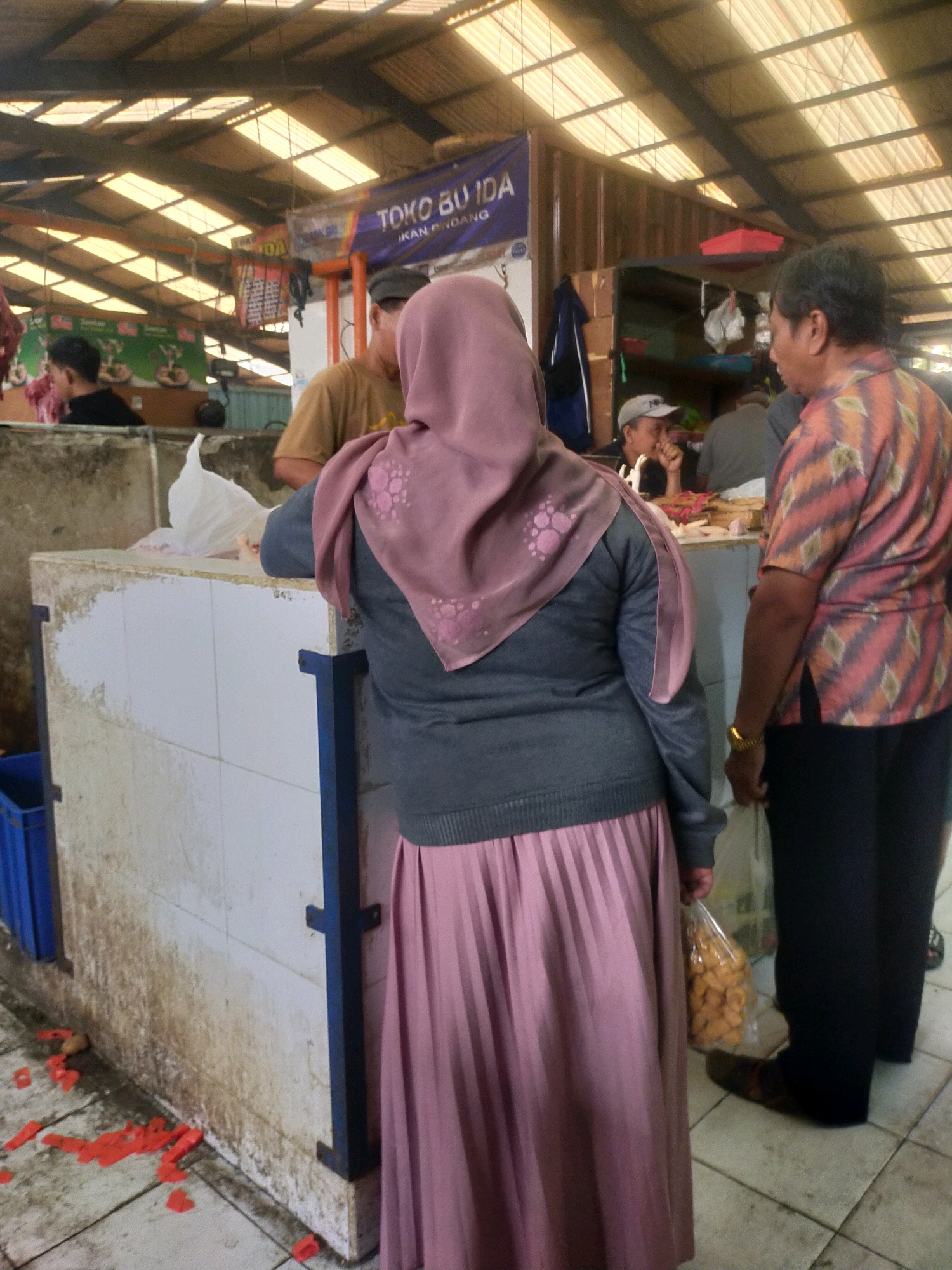 In the city of Bululawang, Indonesia, a market scene unfolds beneath a corrugated metal roof with translucent panels that allow natural light to filter through. The foreground features a woman from behind, wearing a mauve hijab, a grey sweater, and a pleated pink skirt. She is standing at a white tiled counter, with a plastic bag containing light-colored snacks in her right hand. To her right, a man in a patterned orange and black shirt and black pants stands facing away from the camera, holding a white plastic bag. Behind the counter, a man wearing a beige t-shirt and a grey baseball cap is visible. The market floor is tiled and appears slightly damp, with scattered red plastic items on the ground. In the background, a banner with Indonesian text, "TOKO BUIDA IKAN PONDANG," is displayed. Other market stalls and shoppers are faintly visible in the distance.