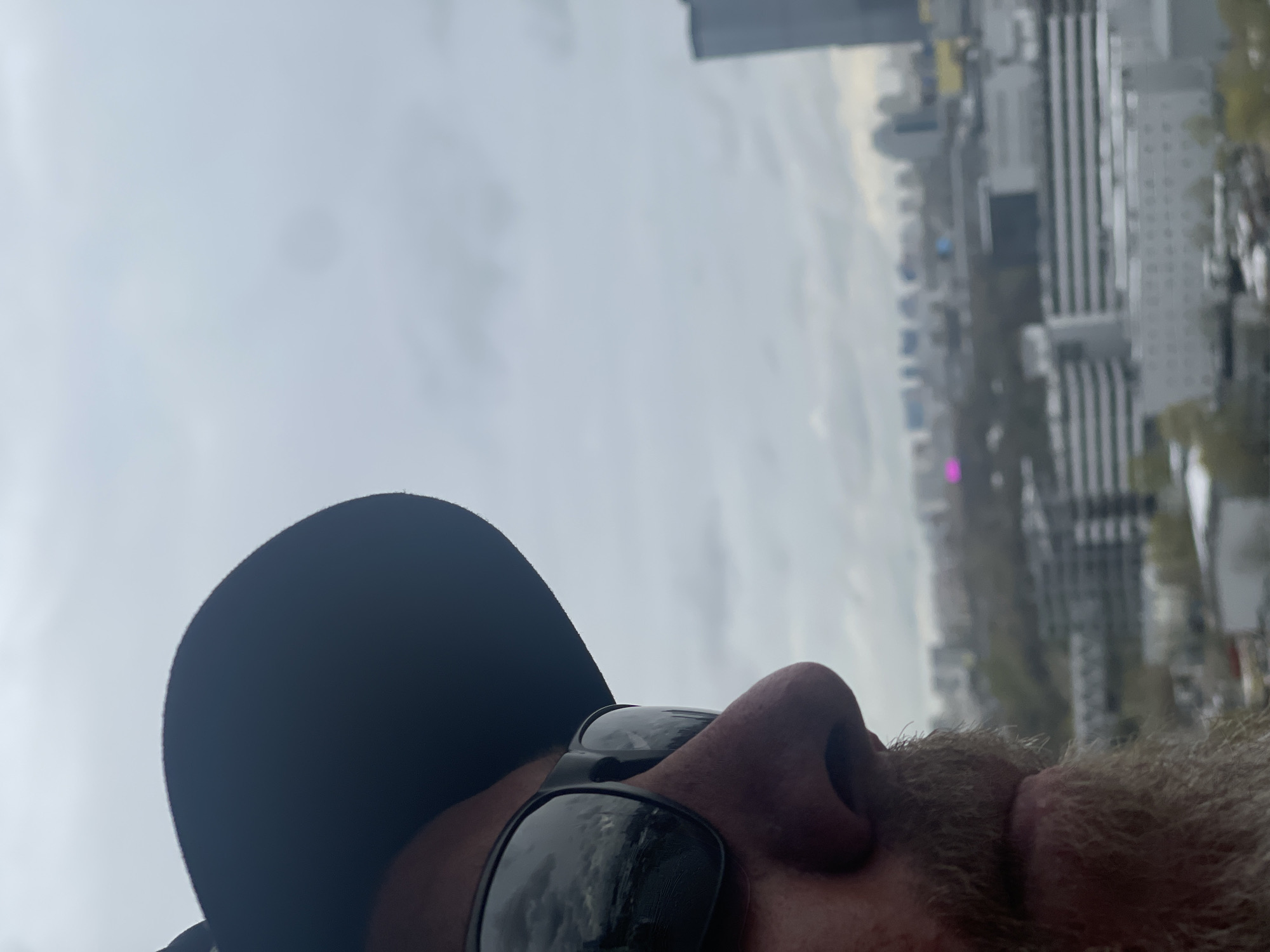 The media file displays a close-up, side profile of a person, likely male due to visible stubble or a short beard, wearing a dark baseball cap and black sunglasses. The person occupies the foreground, looking towards a city skyline in the background. The image is oriented vertically, with the person's head positioned at the bottom, looking towards the right side of the frame.
In the background, an urban landscape unfolds under an overcast, light gray sky. Numerous buildings of varying heights and architectural styles constitute the city skyline, suggesting a modern environment like Khimki, Russia. Some buildings feature prominent vertical lines, and a distinctive bright pink light or sign can be seen in the middle distance among the structures. Patches of trees or foliage are visible on the right side of the background, breaking up the concrete expanse.
The time of day appears to be daytime, with soft, diffused lighting consistent with an overcast or cloudy sky. There is no direct sunlight visible. The individual's expression is not discernible due to the sunglasses and partial view, but they appear to be observing the city. No specific activity beyond viewing the landscape is evident. There is no visible text in the image.