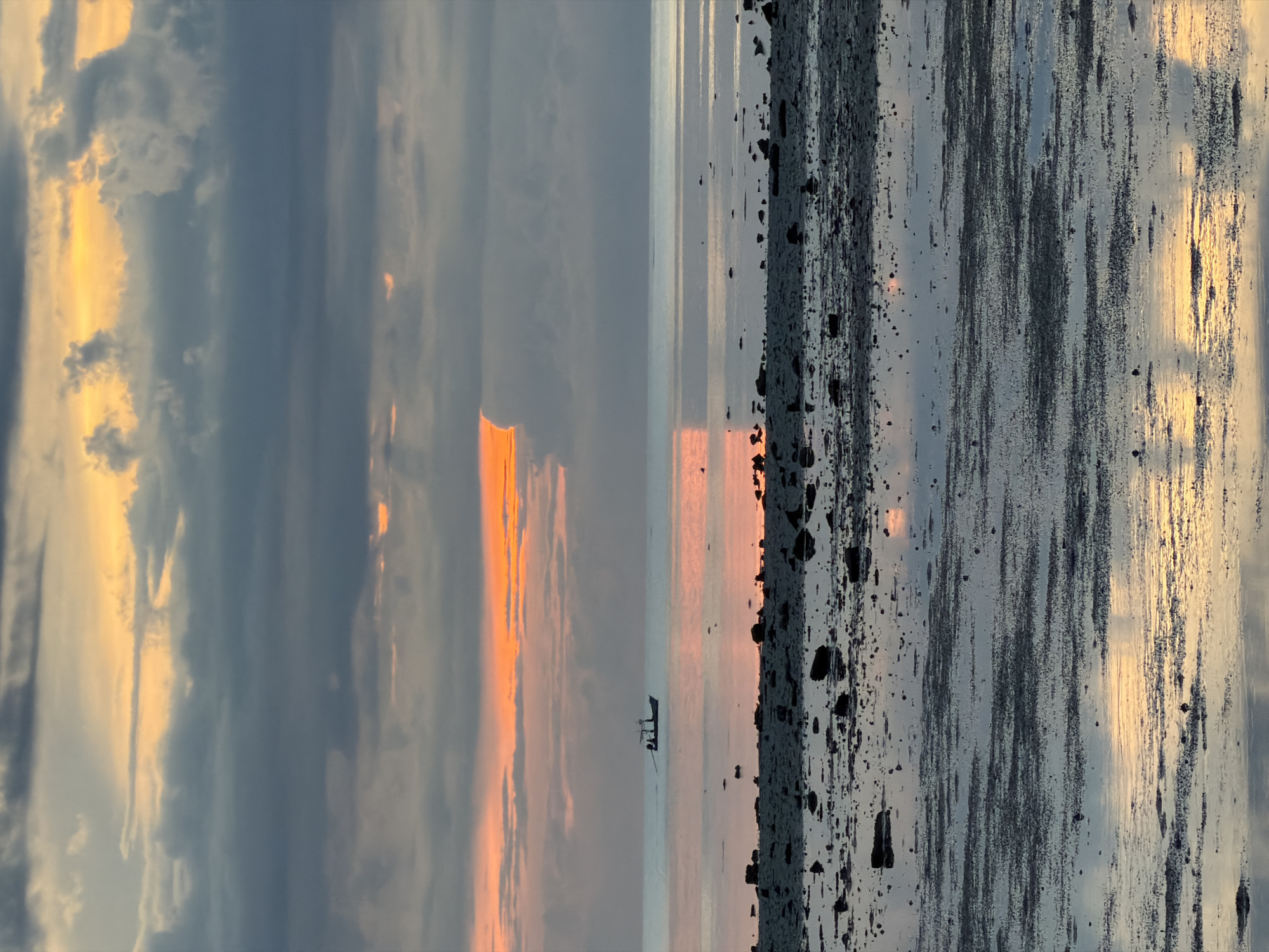 The image depicts a serene coastal scene at Baan Plai Laem, Thailand, during either sunrise or sunset. The sky is filled with dramatic, layered clouds tinged with orange and yellow hues from the low-lying sun, contrasting with darker, grey clouds. A narrow band of bright orange light pierces through a gap in the clouds just above the horizon.
The ocean stretches out, calm and reflective, mirroring the colors of the sky. In the distance, a small fishing boat with a silhouetted figure or figures on board is visible. The foreground consists of a rocky, wet shore with scattered stones and pebbles, reflecting the ambient light and the colors of the sky. The overall impression is one of tranquility and natural beauty.