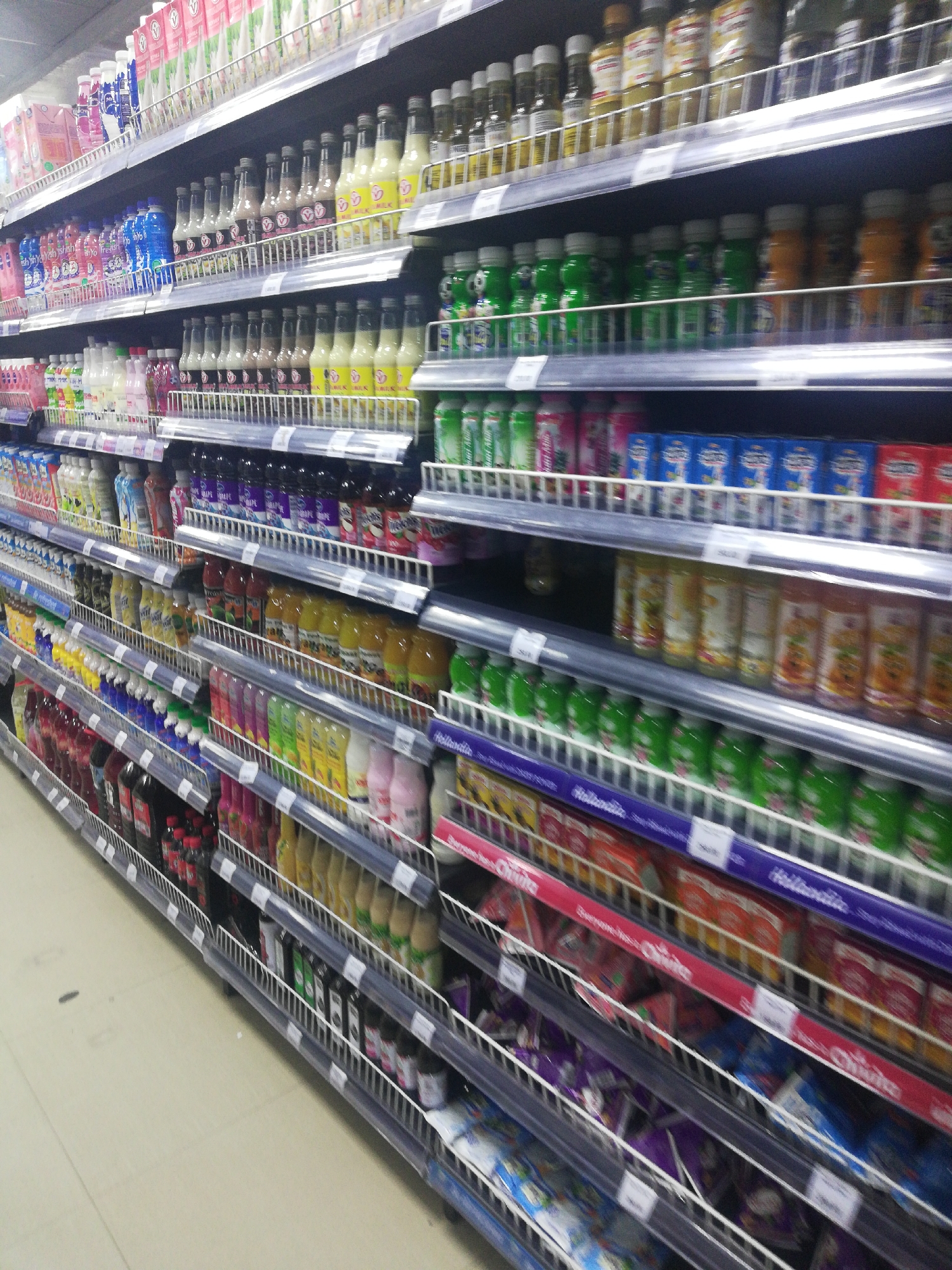 The image shows supermarket shelves stocked with various beverages and drinks. The shelves are filled with bottles and cans, arranged by type and brand. The scene appears to be inside a retail store, likely a supermarket or convenience store. While it is difficult to confirm the location definitively, the types of products and overall aesthetic suggest it could be in Lagos, Nigeria. The shelving units are metallic and display pricing labels. The floor is a light-colored tile.