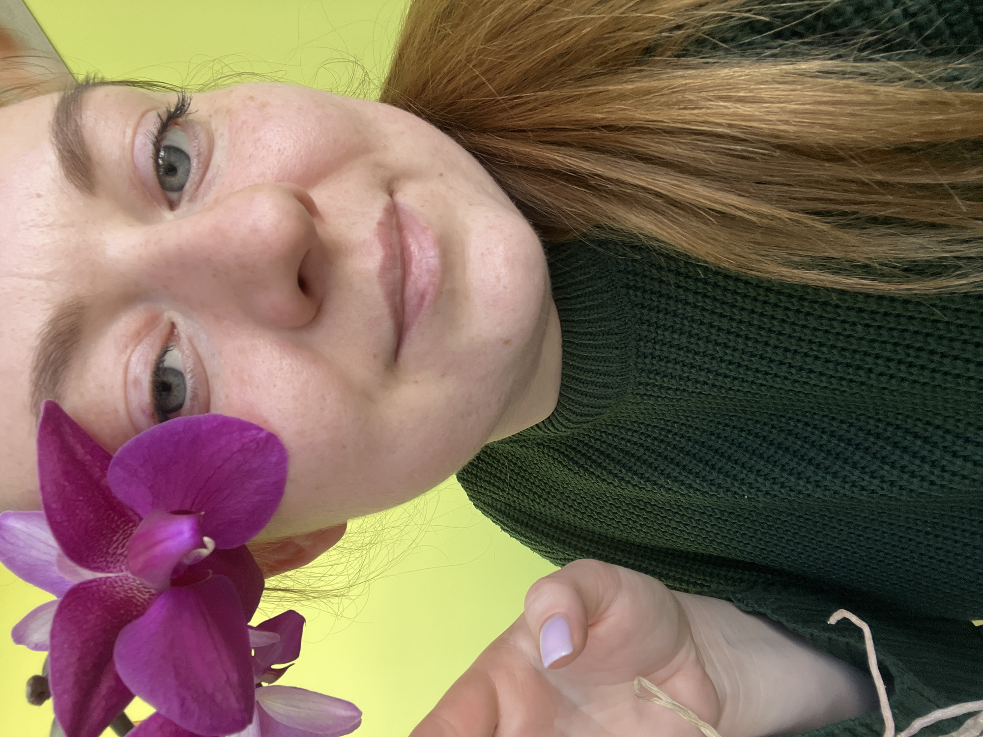 A fair-skinned woman with red-blonde hair is angled towards the camera, wearing a green sweater. A purple orchid is in the lower left of the frame. The background is a solid yellow. The scene does not appear to be in Klin, Russia.