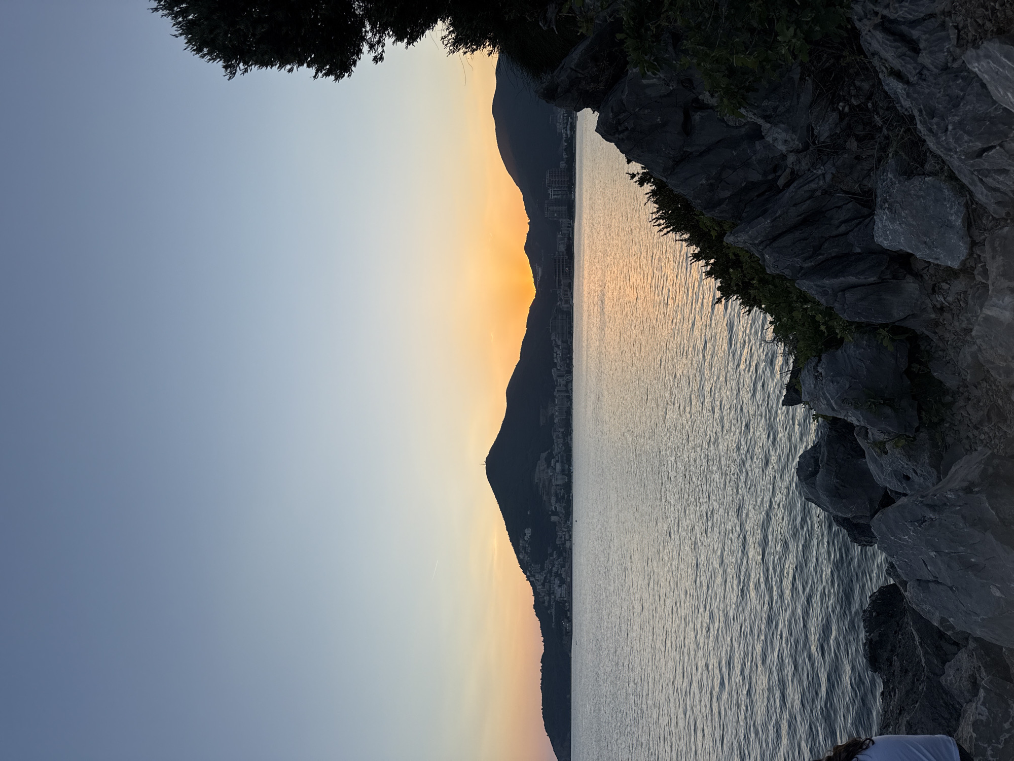 A wide-angle, vertical, low-light shot captures a sunset over the sea, with the city of Budva, Montenegro, nestled on a hillside in the background. The sky transitions from a pale blue at the top to a soft orange and yellow near the horizon where the sun is setting between two dark, silhouetted mountains. The sea is calm, reflecting the sky's colors, and ripples are visible. In the foreground, dark, jagged rocks and green shrubbery create a natural, rugged border along the coastline. A portion of a person, wearing a white shirt, is visible on the left side of the frame, sitting on the rocks. The overall atmosphere is serene and tranquil.