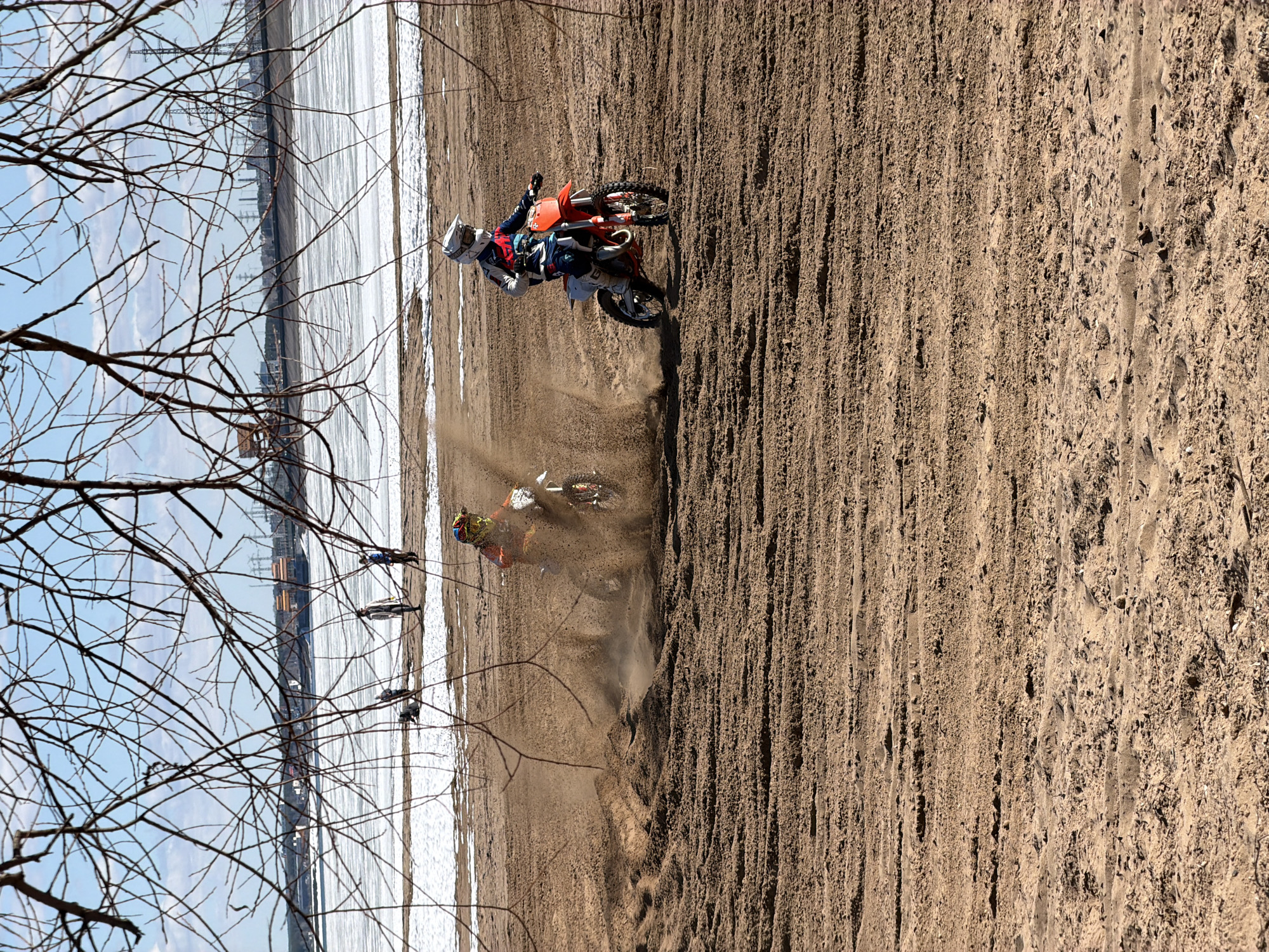 Two people on motorcycles ride on a sandy beach next to a body of water, likely the Kama River, in Chaikovsky, Russia. Branches of bare trees are in the left foreground. Snow is present along the edge of the water. Power lines and buildings are visible in the background across the water. The dirt bike in the foreground is kicking up a cloud of dust.