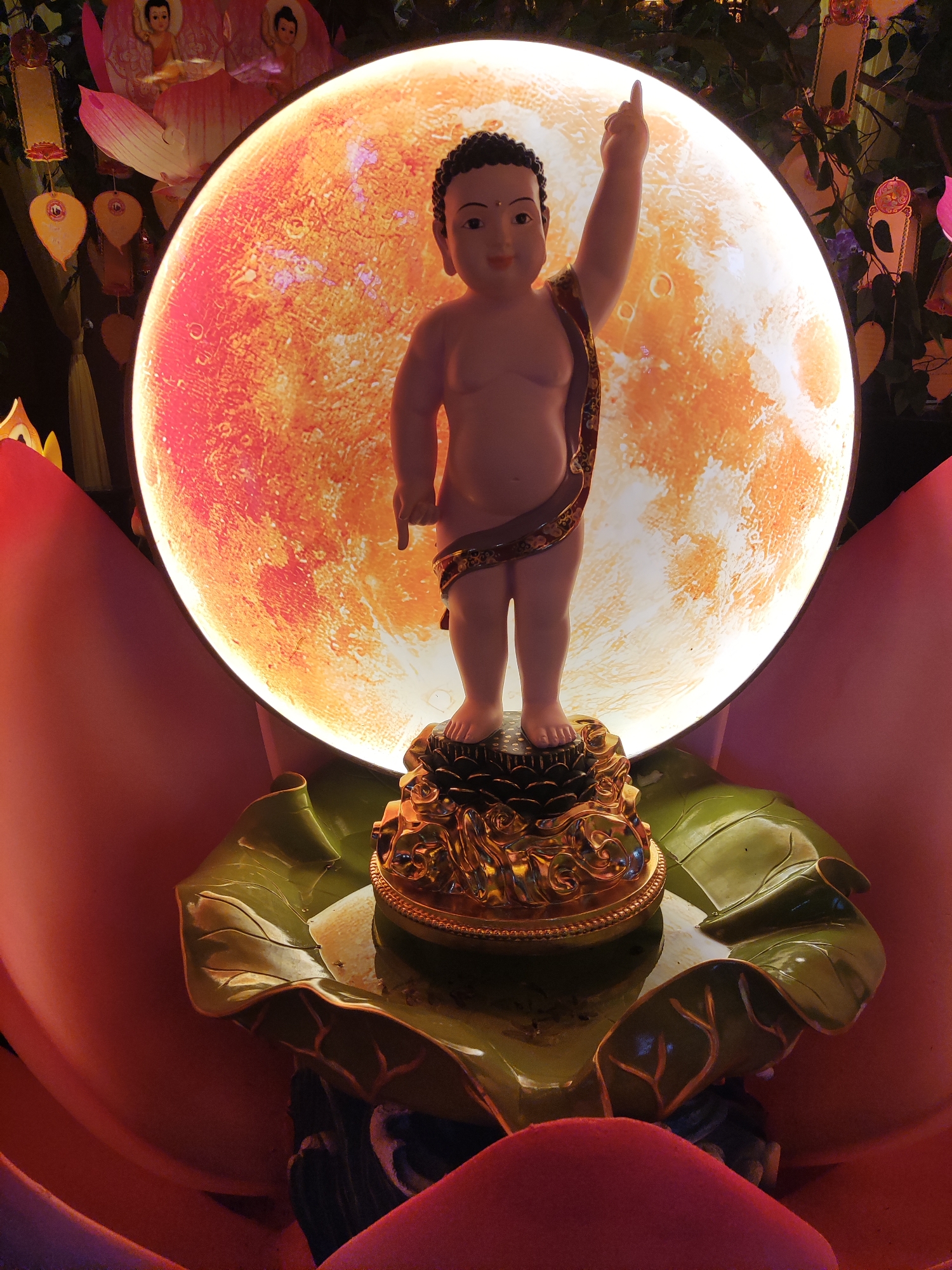 A serene, light-skinned statue of the infant Buddha is the focal point of this vertically oriented image. The baby Buddha, with its right arm raised and index finger pointing skyward and its left arm angled downwards with index finger pointing to the earth, stands on an elaborate, golden, multi-tiered pedestal. This pedestal rests upon a large, glossy green lotus leaf, which itself is nestled within vibrant, open pink and red lotus petals forming the base of the display.
Directly behind the statue, a large, glowing circular disc dominates the background, radiating a warm, textured orange-red light, resembling a stylized full moon or celestial body with visible crater-like markings. The illumination from this disc casts a radiant glow on the scene. In the periphery of the dark background, subtle decorative elements are visible, including small Buddha figures depicted on pink lotus blossoms and numerous golden, heart-shaped leaf ornaments, likely symbolizing Bodhi leaves.
The overall setting is brightly lit, indicating an indoor or evening/nighttime devotional display. The statue's iconic pose, with fingers pointing up and down, is a symbolic representation of the infant Buddha at birth, signifying "heaven above, earth below, I alone am the World Honored One," a common motif in Buddhist celebrations like Vesak or Hanamatsuri. There is no visible text.