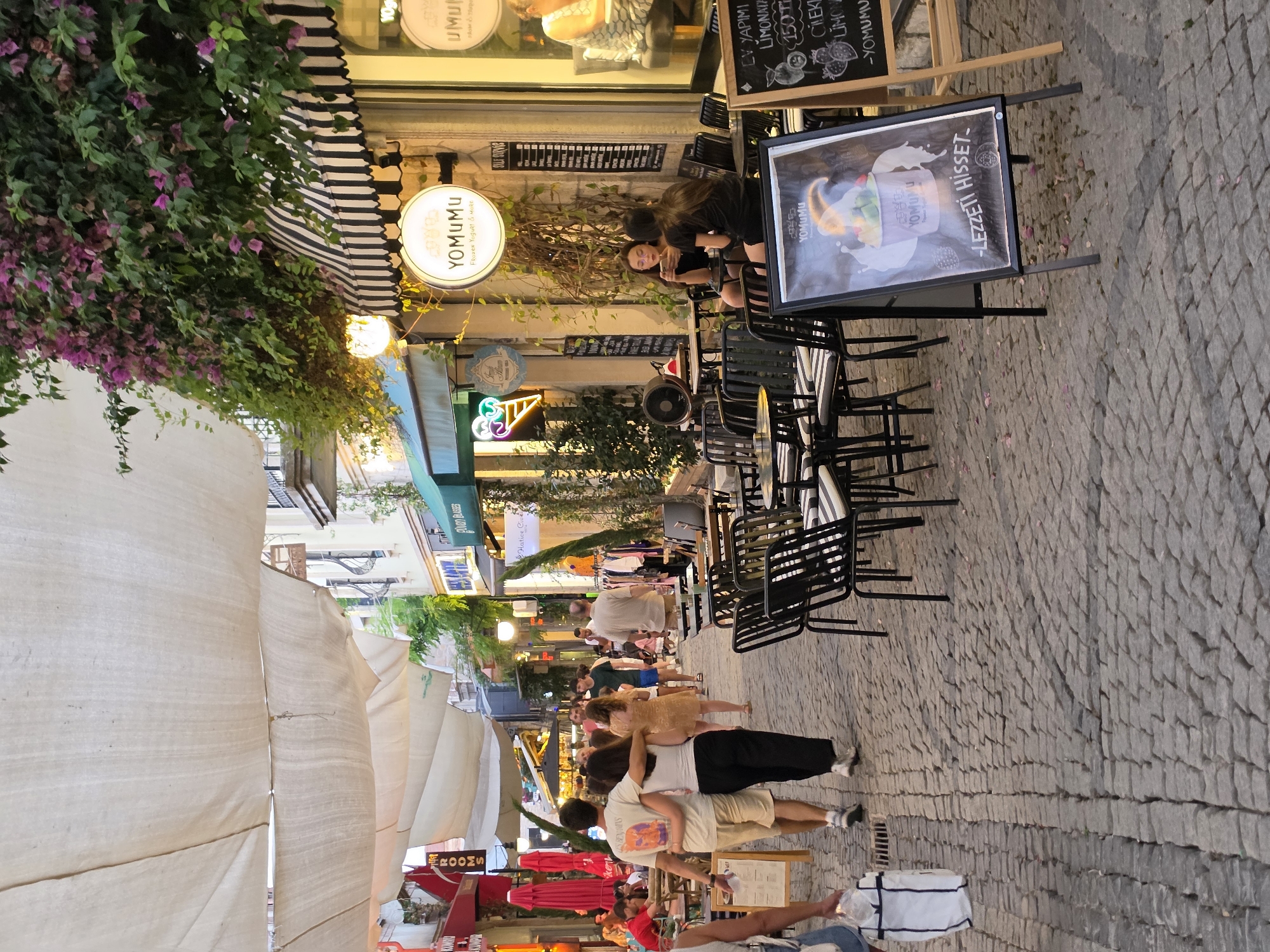 This image captures a street scene in Urla, Turkey, likely during the evening, given the lighting and atmosphere. The cobblestone street is lined with shops and cafes, creating a lively pedestrian area.
In the foreground, two people walk away from the camera, with one person's arm around the other. To the right, outdoor seating areas for cafes are visible, with empty chairs and tables. One cafe, "YOMUMU," is prominently featured with its distinctive black and white striped awning and a circular sign. A menu board outside the cafe displays an image of a dessert and text, including "LEZZETI HISSET." Inside the cafe, two women are seated at a table, engaged in conversation.
Further down the street, more shops and cafes are visible, with awnings providing shade and colorful signs indicating businesses. People are seen strolling along the street and seated at outdoor tables. Lush green plants and blooming purple flowers cascade from above, adding to the charm of the setting. The overall impression is that of a vibrant and popular urban destination, likely a tourist spot in Urla.