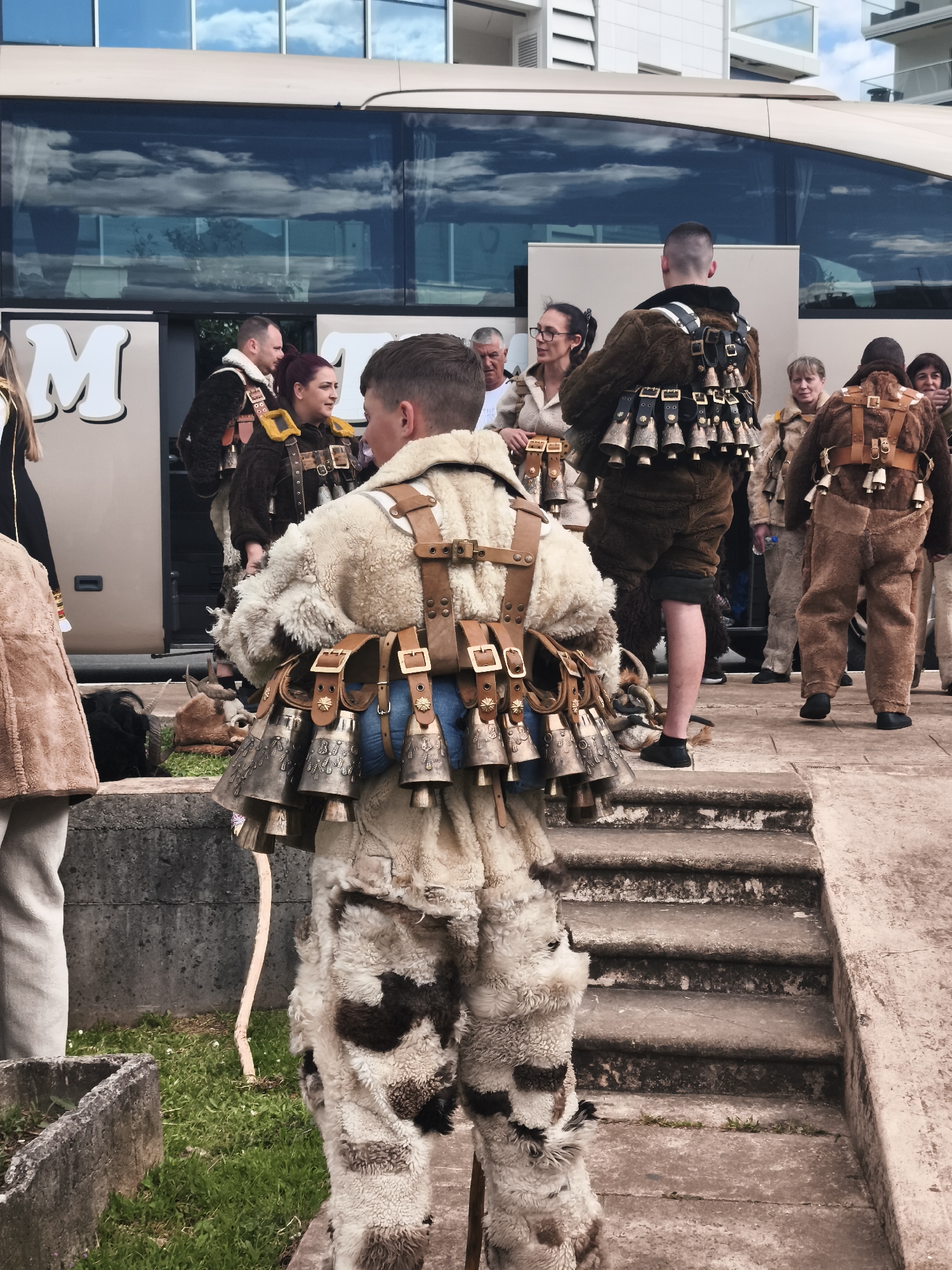 A medium shot depicts a group of people dressed in traditional costumes with bells attached to their clothing standing on a concrete surface. The individual in the foreground is wearing a light-colored, furry suit with brown and white spots, and a leather harness holding several large metal bells. The suit is designed to mimic the appearance of animal fur. Other individuals are dressed in similar attire but with varying colors and fur types.
The group is positioned in front of a bus with large reflective windows that display the sky and clouds. Behind the bus is a modern building with a white facade and multiple floors. The setting appears to be in an urban area, as evidenced by the concrete steps and surfaces, as well as the presence of the bus and modern architecture. A small patch of grass and a concrete planter are visible in the lower left corner of the image. The people seem to be gathered for an event or performance, and their traditional clothing suggests a cultural or festive occasion.