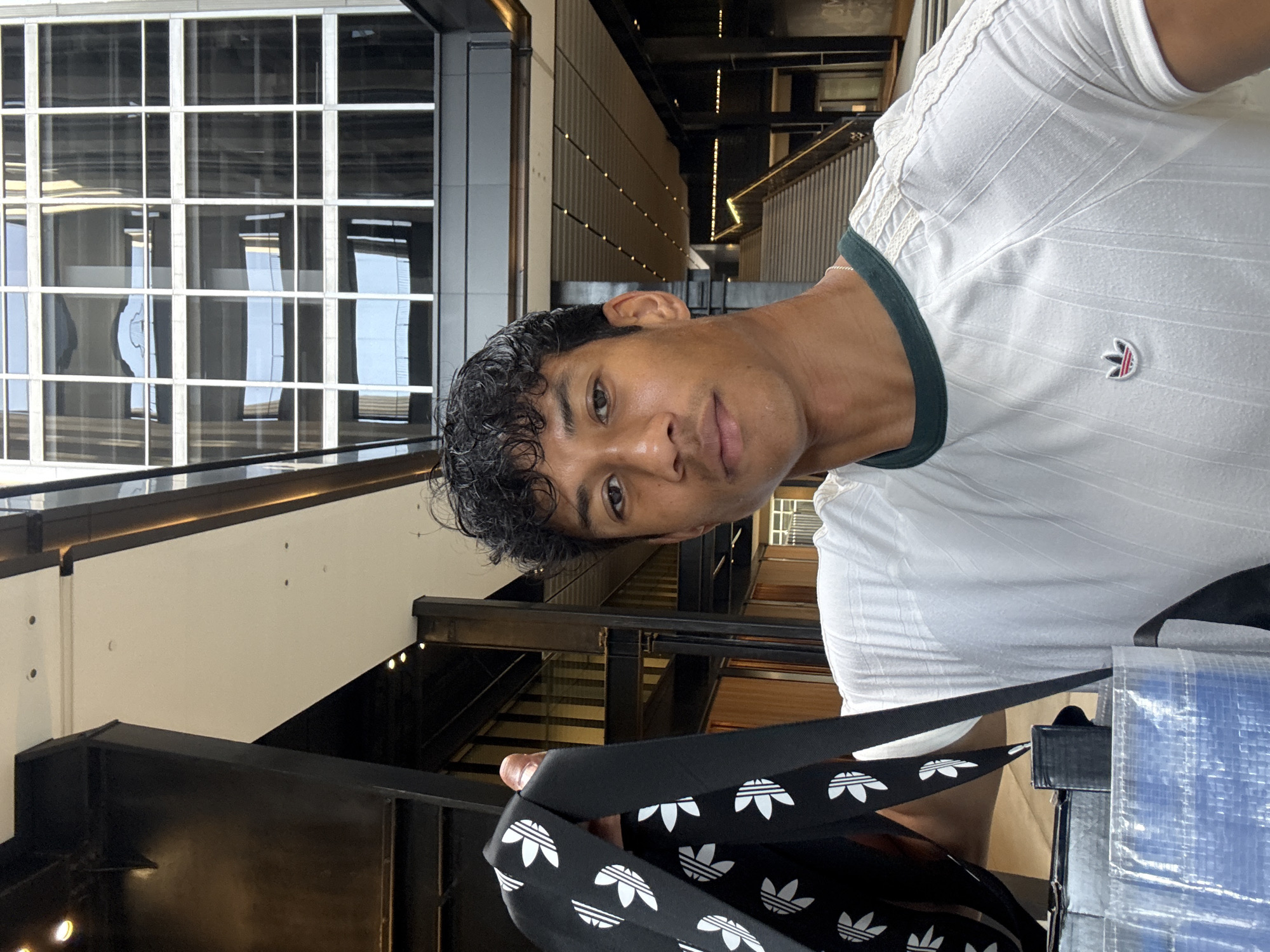 A young adult male with short, curly, black hair and tan skin is looking at the camera. He is wearing a white Adidas t-shirt with a green trim. He is holding a black Adidas-patterned strap, presumably of a bag. The photograph is taken in a building with large window panels and complex interior architecture featuring multiple levels and metal railings. The location is within the city of Los Angeles, United States of America.
