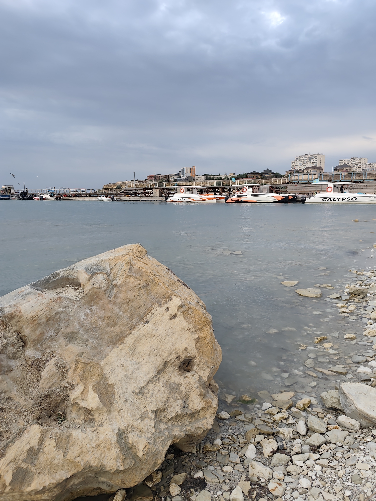 The image depicts a coastline in Aktau, Kazakhstan, likely near a marina or harbor. In the foreground, a large, light-colored boulder dominates the left side of the frame, with a rocky shore leading into the water on the right. The water is calm and reflects the overcast sky.
In the middle ground, several boats are docked or moored along a pier. Some of these boats are leisure vessels with names visible, such as "CALYPSO" and "NEMO". Beyond the pier, a series of buildings, possibly residential or commercial, are visible along the elevated coastline, with some structures appearing to be under construction. The sky is heavily clouded, suggesting a subdued or approaching inclement weather. A few seagulls are in flight near the pier. The overall scene is one of a coastal urban environment under a cloudy sky.