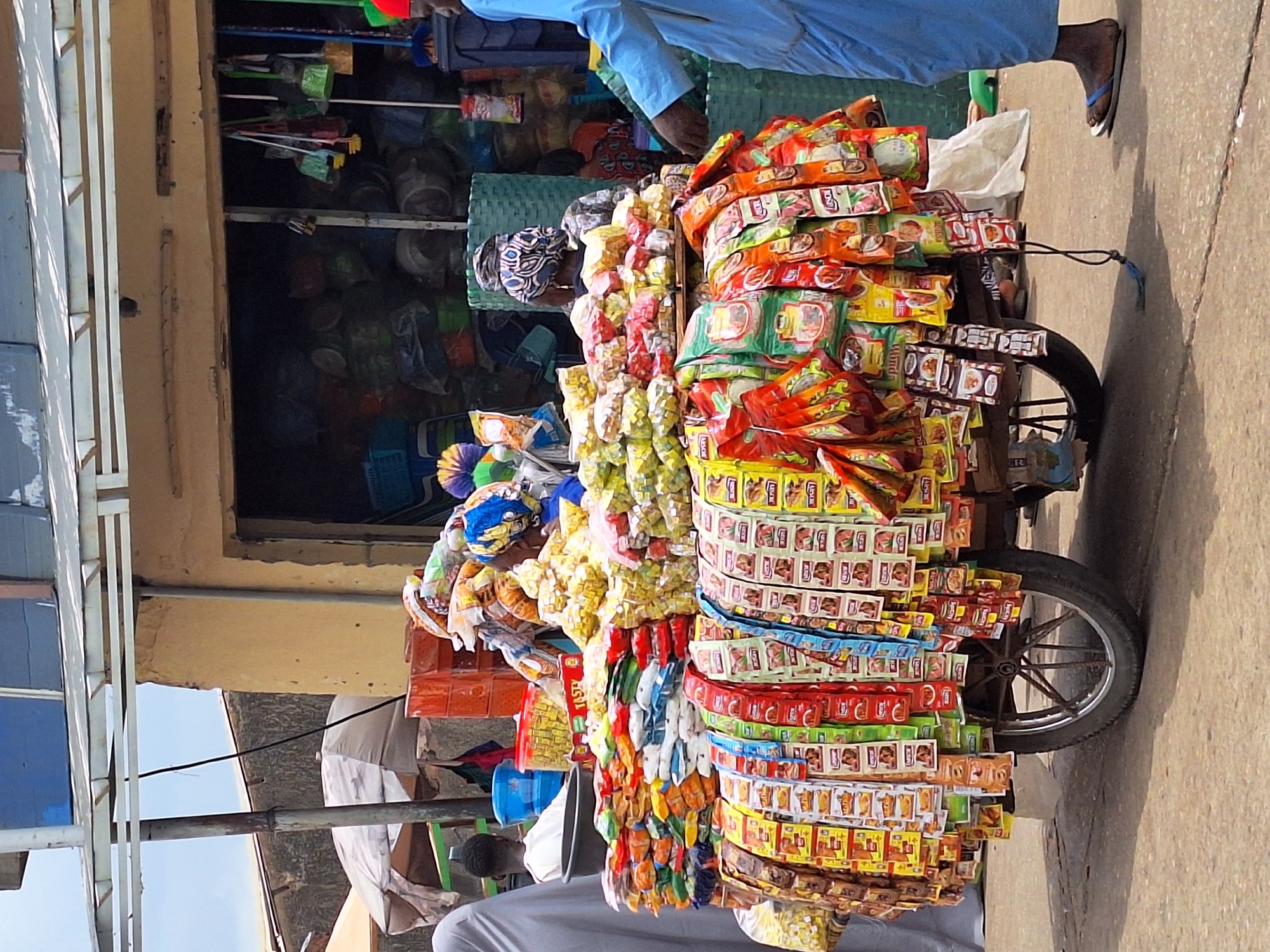 This image captures a vibrant street market scene in Maiburiji, Nigeria, during the daytime. The central subject is a brightly decorated two-wheeled vendor's cart, meticulously laden with a vast array of small, packaged goods.
The cart is overflowing with colorful sachets and packets, primarily displaying what appear to be snacks, instant food items, or spices. These packages are arranged in dense vertical rows and bundles, maximizing the display area. Notable brands visible on some packets include "LAY'S" (chips), "MAMA GOLD" (a common instant noodle brand), "P.K.", and "TASTY". The bright reds, yellows, oranges, and greens of the packaging create a striking visual impact. A black cord, possibly for steering or braking, is visible attached to the cart near its wheel.
A person, likely the vendor, is partially visible to the right of the cart, wearing a light blue long-sleeved garment and dark flip-flops. Their feet and lower legs are visible on the concrete ground, casting a shadow. Another person is partially visible sitting or crouching below the cart, wearing a light grey top.
In the background, an open-fronted shop or stall is visible, stocked with various household items. One can discern brooms, brushes, woven baskets, and other containers, indicating a general store setting. The building itself is a simple structure with a light brown wall. On the far left, a metal framework, possibly supporting an awning or another part of a building, is visible against a bright sky.
The scene is illuminated by clear daylight, casting distinct shadows on the concrete ground, suggesting sunny weather. The overall impression is that of a bustling commercial environment characteristic of a local market in Nigeria.